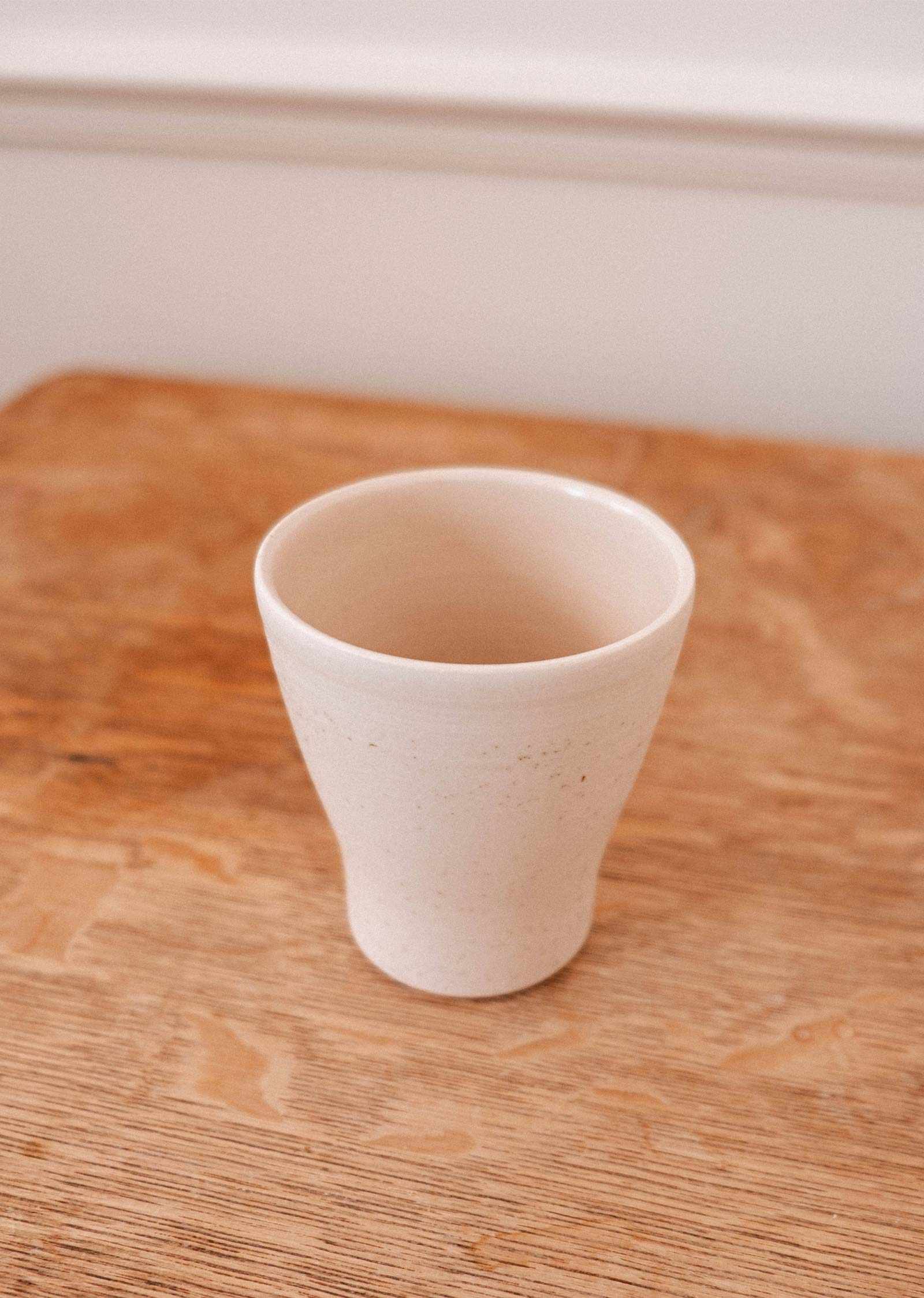 The Small Ceramic Cup by Mimi & August rests on a wooden surface against a light-colored wall.