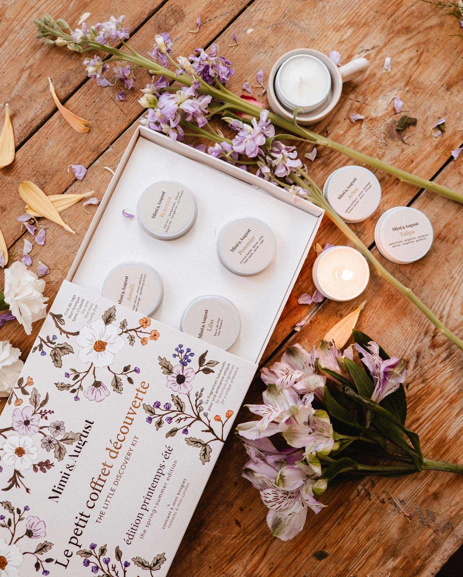 The Spring/Summer Discovery Kit by Mimi & August, featuring five small skincare containers, is displayed open on a wooden table with a mini soy candle, purple and white flowers, loose candles, and flower petals.