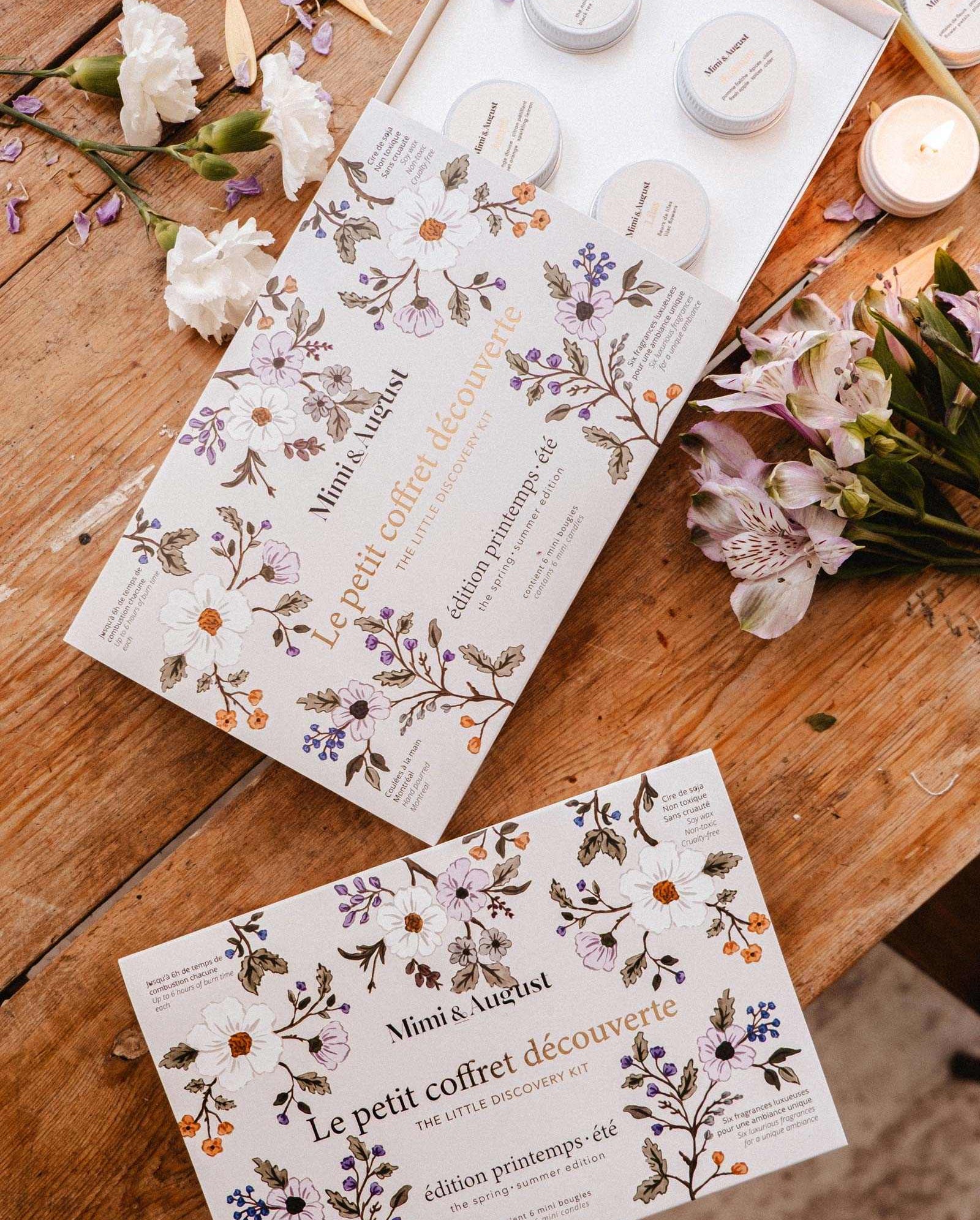 Mimi & August’s The Spring/Summer Discovery Kit and a mini soy candle are displayed on a wooden table with scattered white and purple flowers.