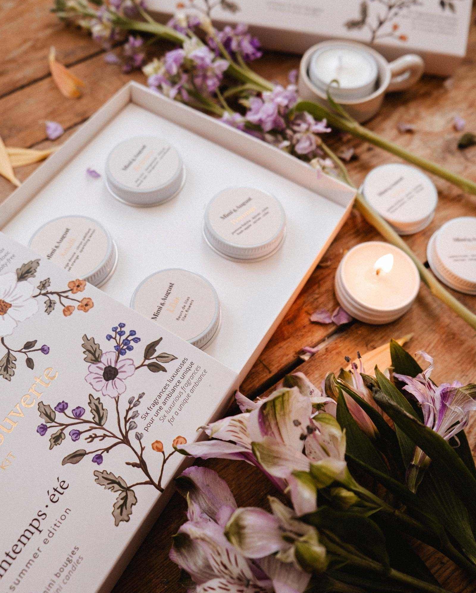 The Spring/Summer Discovery Kit by Mimi & August features an open floral-themed box with small labeled jars and a soy tealight sampler, displayed among flowers, candles, and petals on a wooden surface.