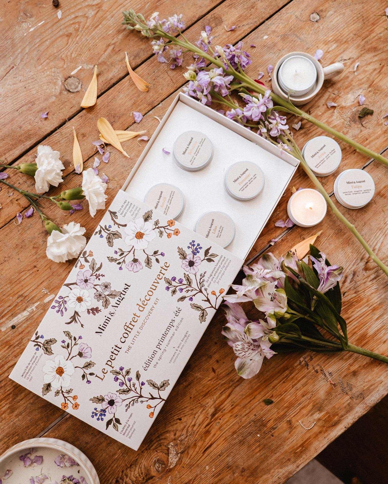 The Spring/Summer Discovery Kit by Mimi & August: an open, floral-themed skincare gift box with small jars, a mini soy candle, and a soy tealight sampler, beautifully arranged among flowers and candles on a rustic wooden table.