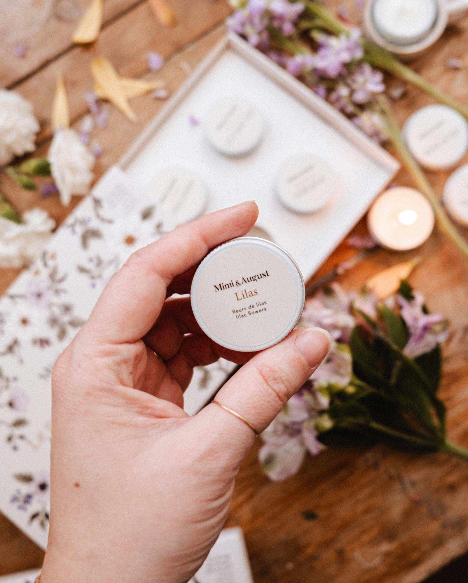 A hand holds a small tin labeled "Lilac" from Mimi & August's Spring/Summer Discovery Kit, displayed with floral-themed items and a mini soy candle—all beautifully arranged on a wooden surface.