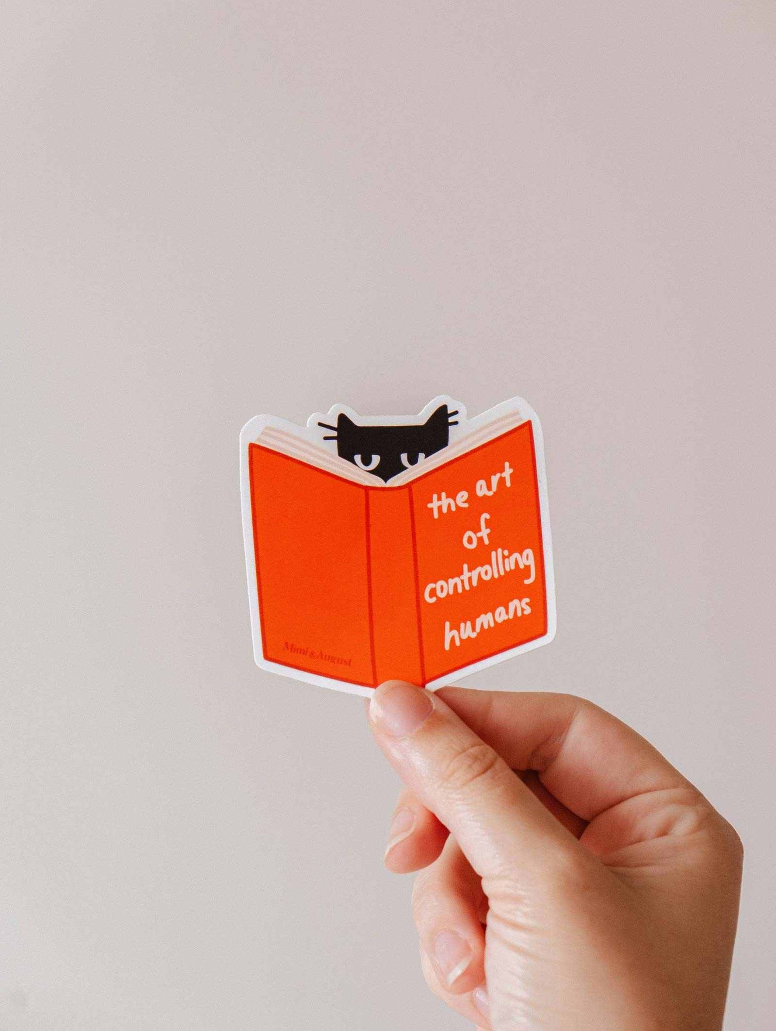 Black cat vinyl sticker titled The Art of Controlling Humans with red book, durable and weather-resistant design