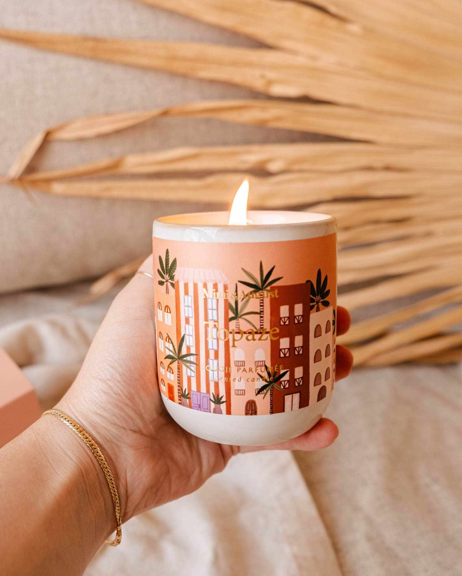 A hand holds a lit Topaze - Reusable Candle by Mimi & August in a white container with an orange and pink design featuring buildings and leaves, creating a tranquil ambience.