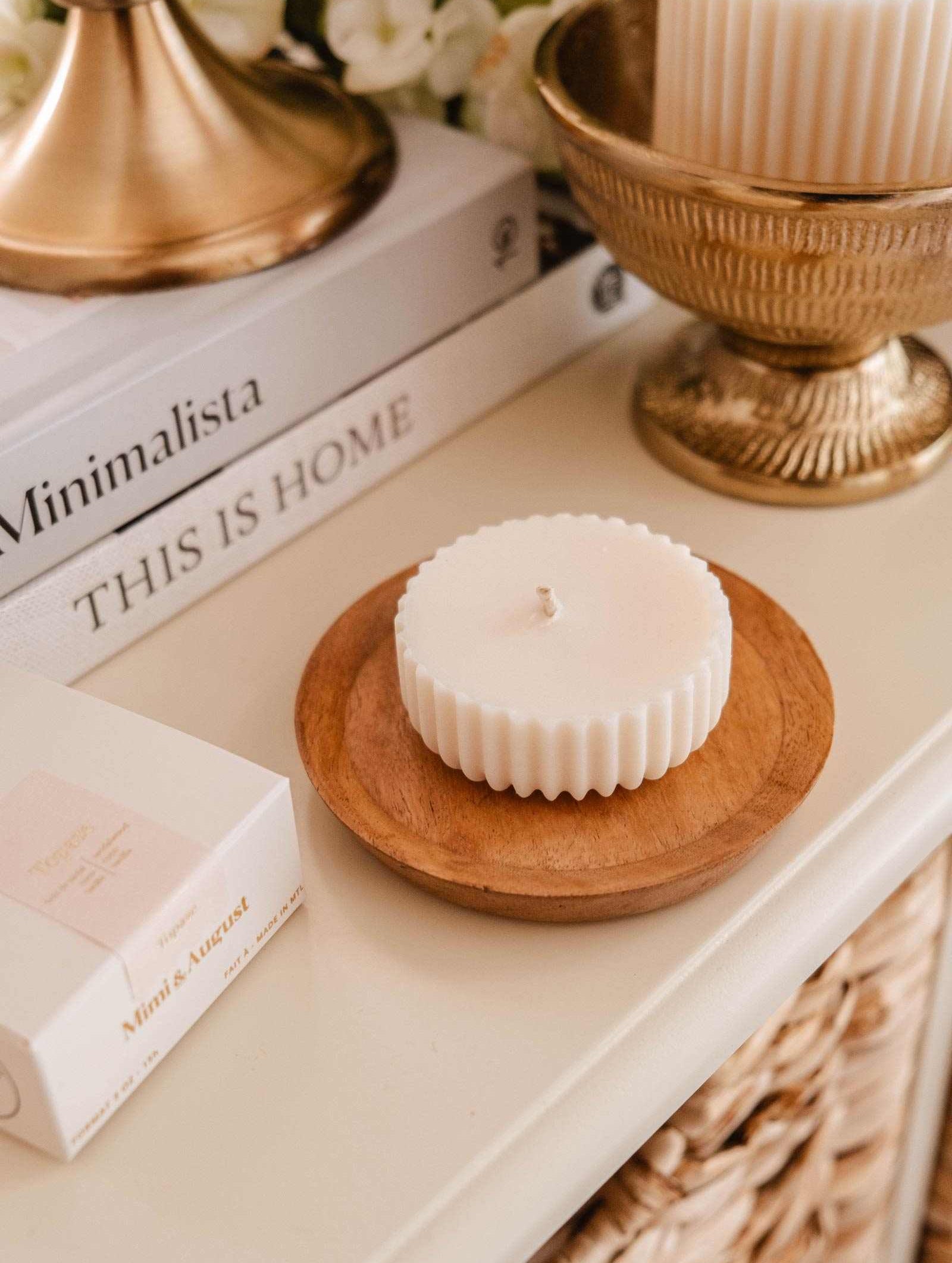 The Candle Refill Cielo by Mimi & August, a white round soy wax refill with a ridged edge, rests on a wooden dish near books, a gold bowl, and a lamp, filling the area with its fig cassis amber scent.