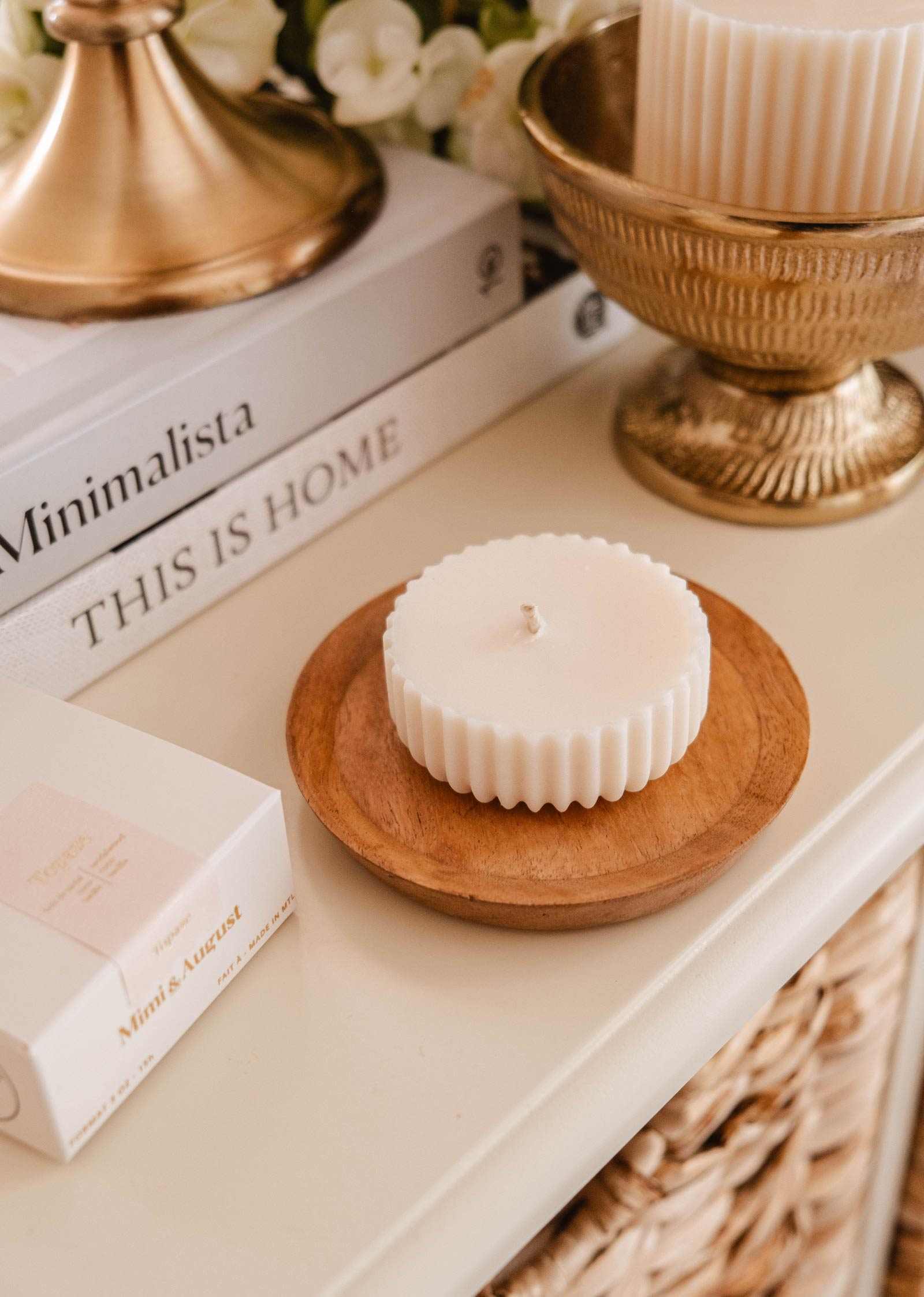 A white, fluted-edge Candle Refill Topaze by Mimi & August sits on a wooden dish near stacked books, a gold bowl, and a "Mini Acust" box, filling the space with sandalwood coconut vanilla warmth.