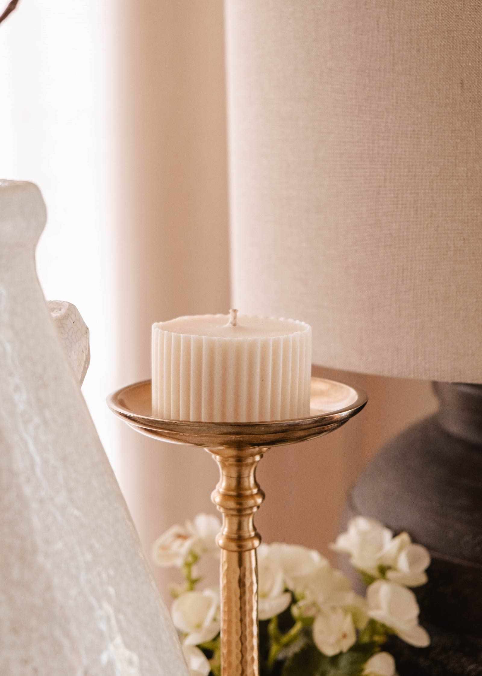 The Candle Refill Topaze by Mimi & August, a white ribbed eco-friendly candle, sits on a gold holder beside a lamp and white flowers, softly illuminating the room with sandalwood coconut vanilla fragrance.