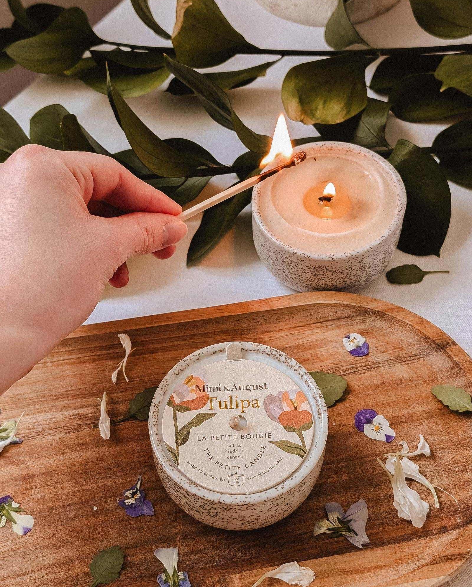 A hand lights the Mimi & August Tulipa - Reusable Candle with wildberries fragrance on a wooden tray next to another candle and scattered flower petals, with green leaves in the background.
