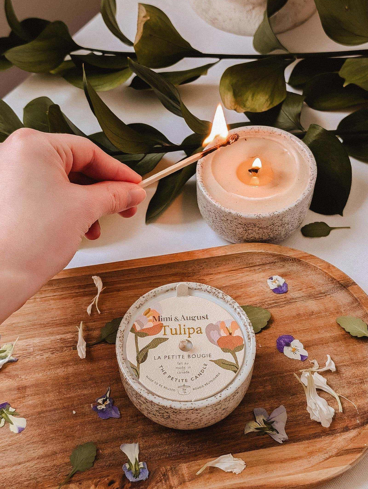 A hand lights the Mimi & August Tulipa - Reusable Candle with wildberries fragrance on a wooden tray next to another candle and scattered flower petals, with green leaves in the background.