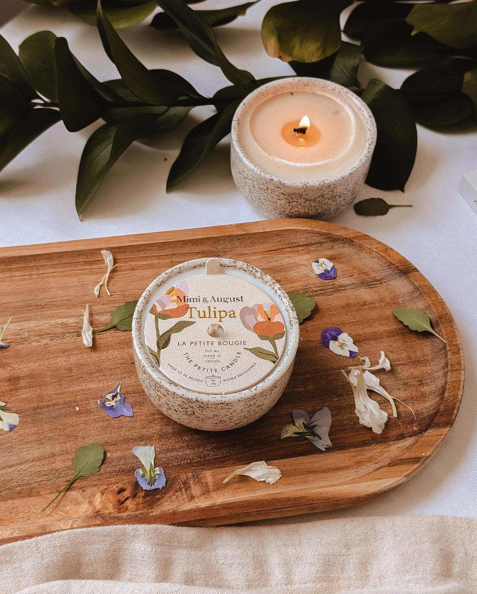 The Tulipa - Reusable Candle by Mimi & August sits round on a wooden board amid scattered petals, with a second lit reusable candle nearby. Greenery in the background enhances the charming scene.