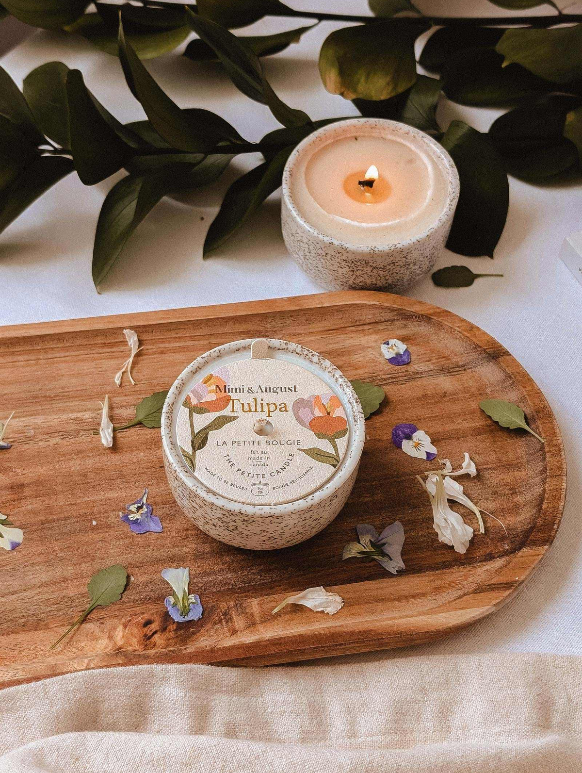 The Tulipa - Reusable Candle by Mimi & August sits round on a wooden board amid scattered petals, with a second lit reusable candle nearby. Greenery in the background enhances the charming scene.