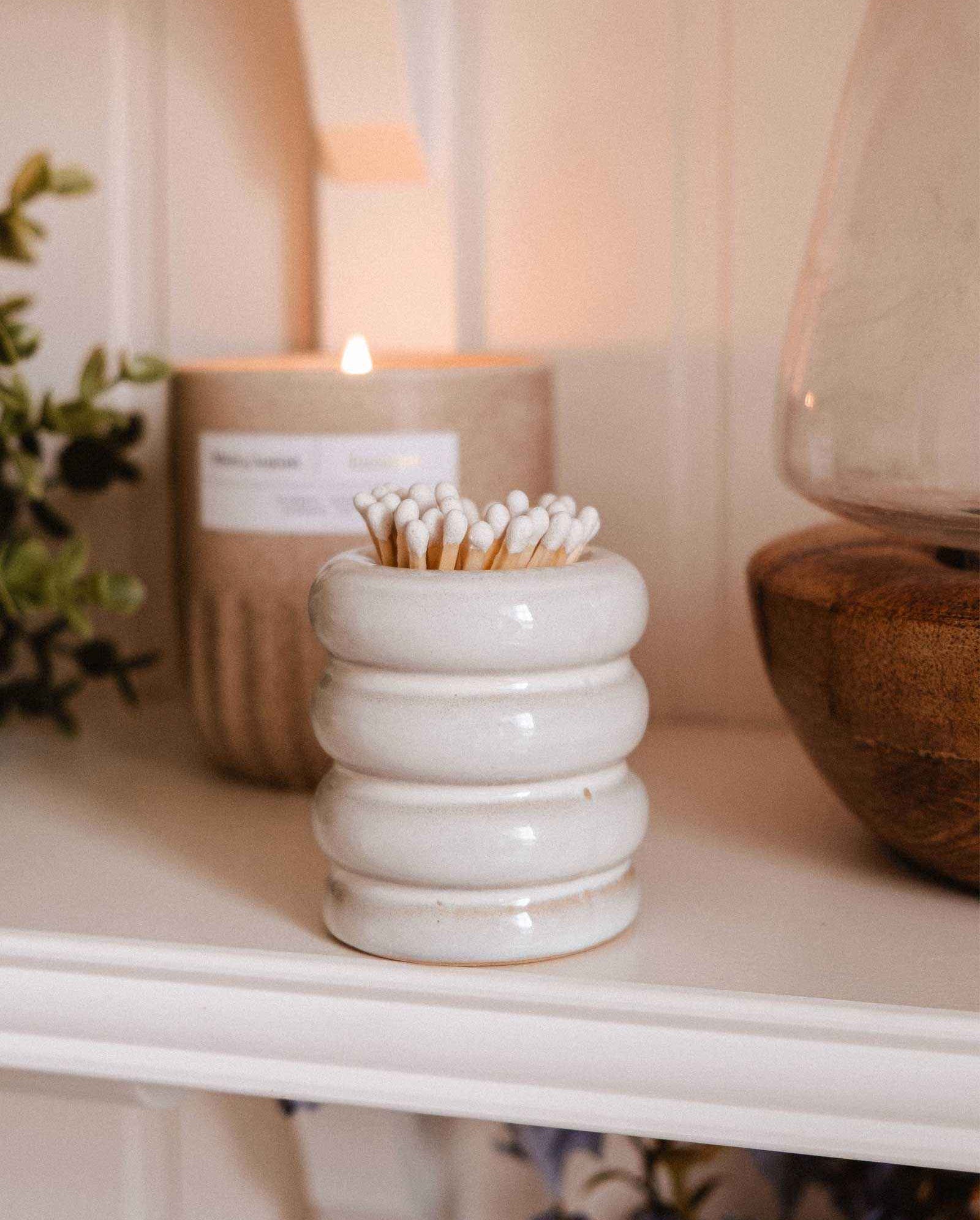 A white Ceramic Match Pot by Mimi & August sits on a shelf next to a lit candle, wooden bowl, cotton swabs in a jar, decorative match holder, and greenery.