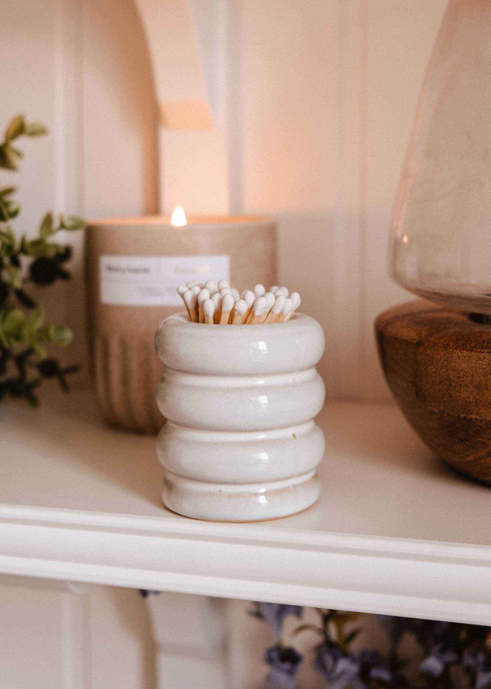 A white Ceramic Match Pot by Mimi & August sits on a shelf next to a lit candle, wooden bowl, cotton swabs in a jar, decorative match holder, and greenery.