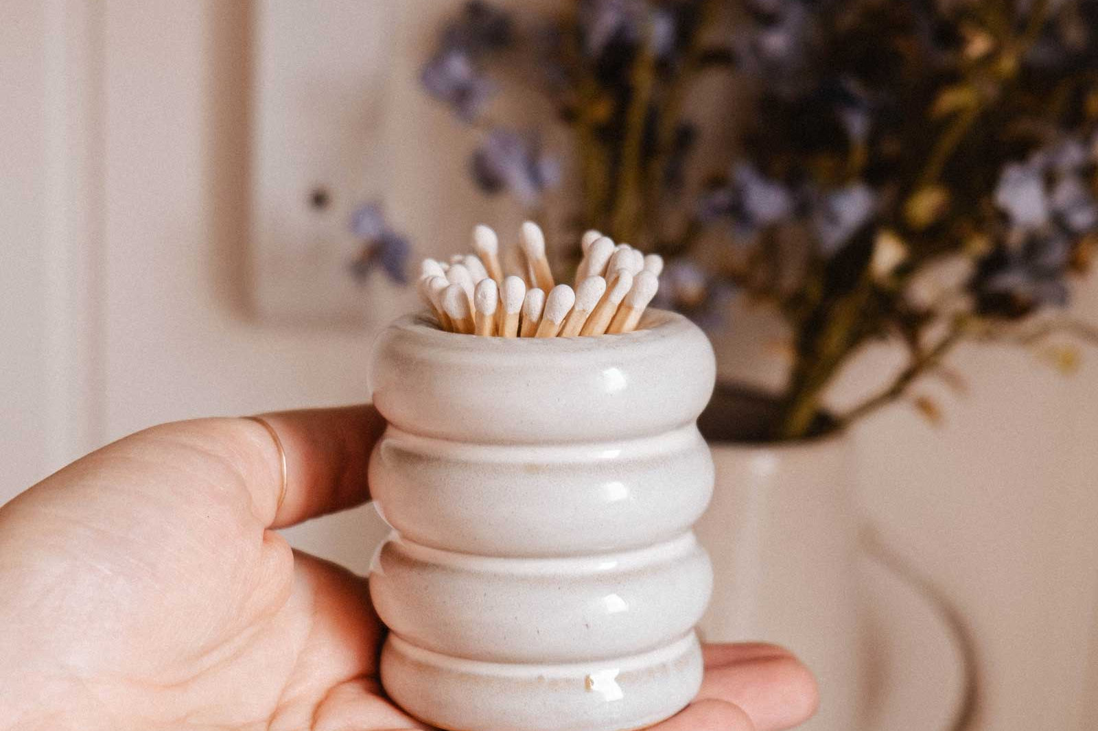 A hand holds the Mimi & August Ceramic Match Pot filled with cotton swabs; blurred purple flowers in a vase appear in the background.