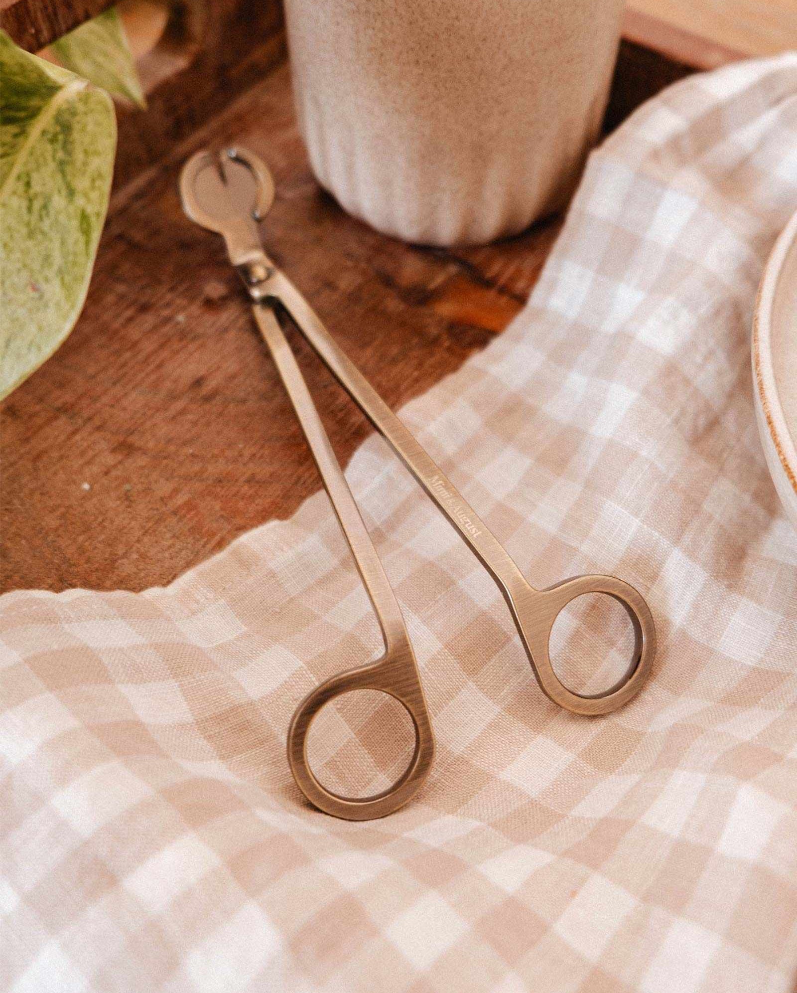 The Mimi & August Wick Trimmer, made of stainless steel, rests on a beige checkered cloth beside a candle and plant, creating an elegant candle accessory for your home.