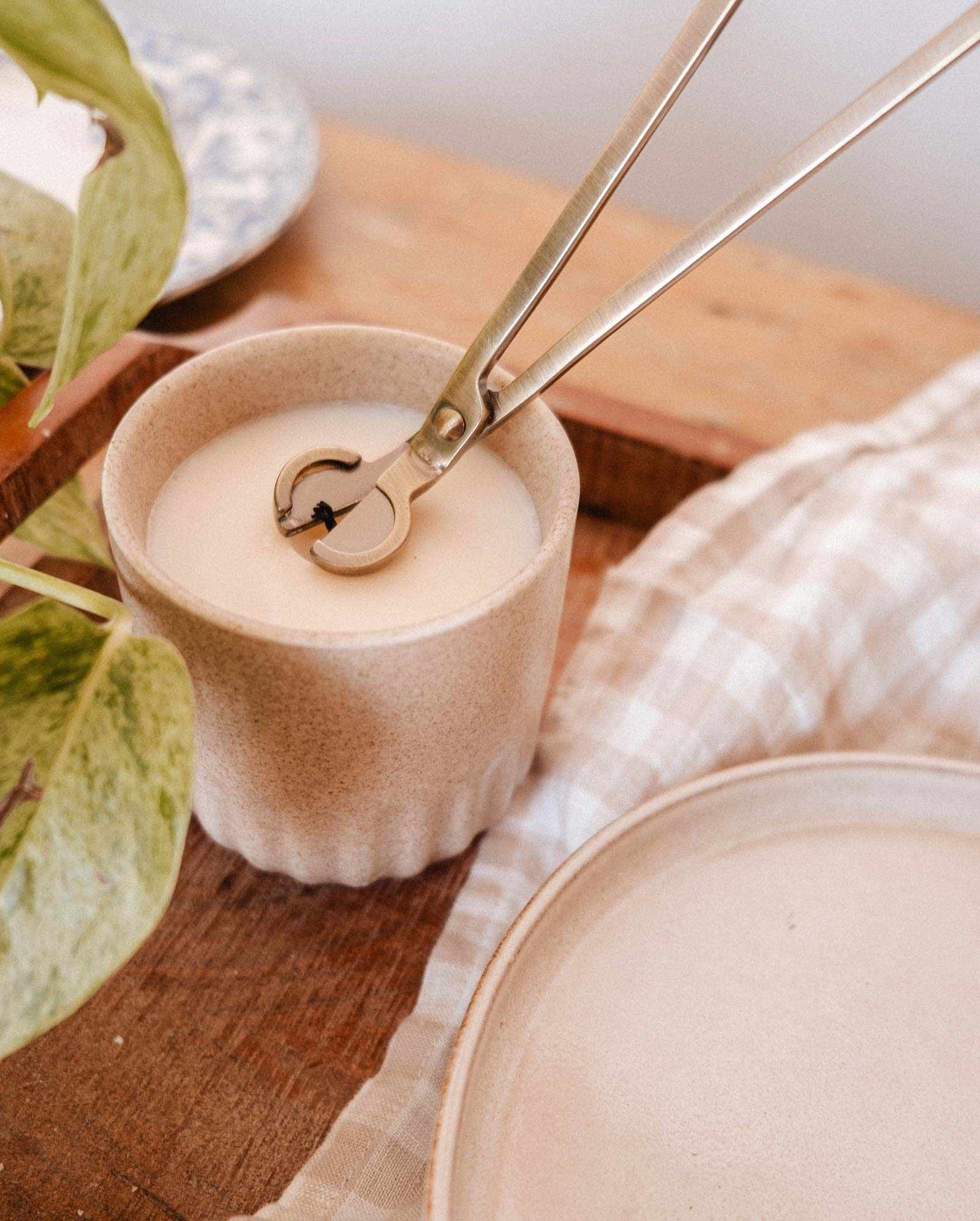 The Mimi & August Wick Trimmer neatly trims candle wicks, shown here with a white candle in a beige ceramic container on a wooden surface—an essential accessory for any cozy space.