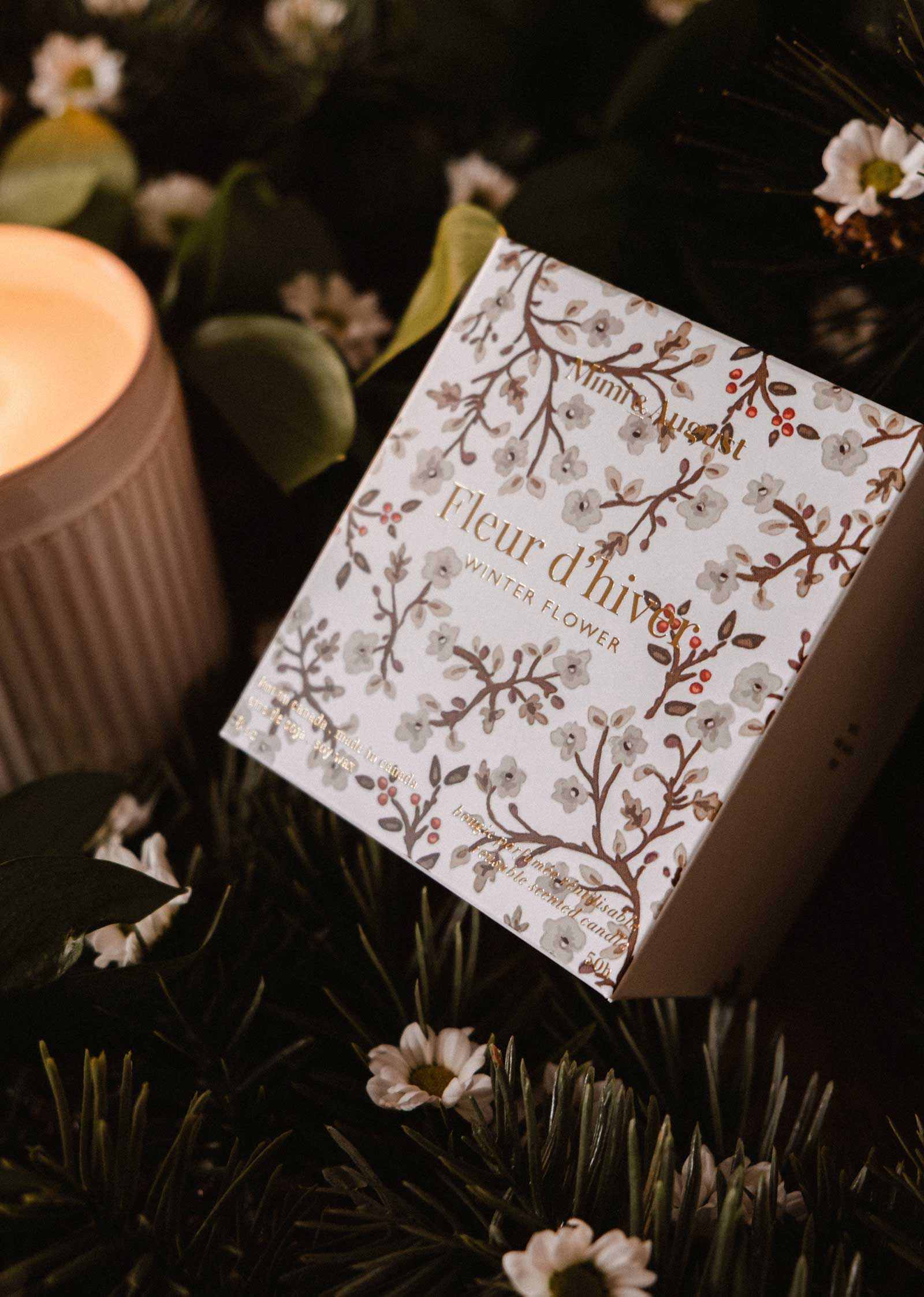 A decorative box labeled "Fleur d’hiver" by Mimi & August sits among greenery and white flowers, beside a glowing Winter Flower - Christmas Candle.