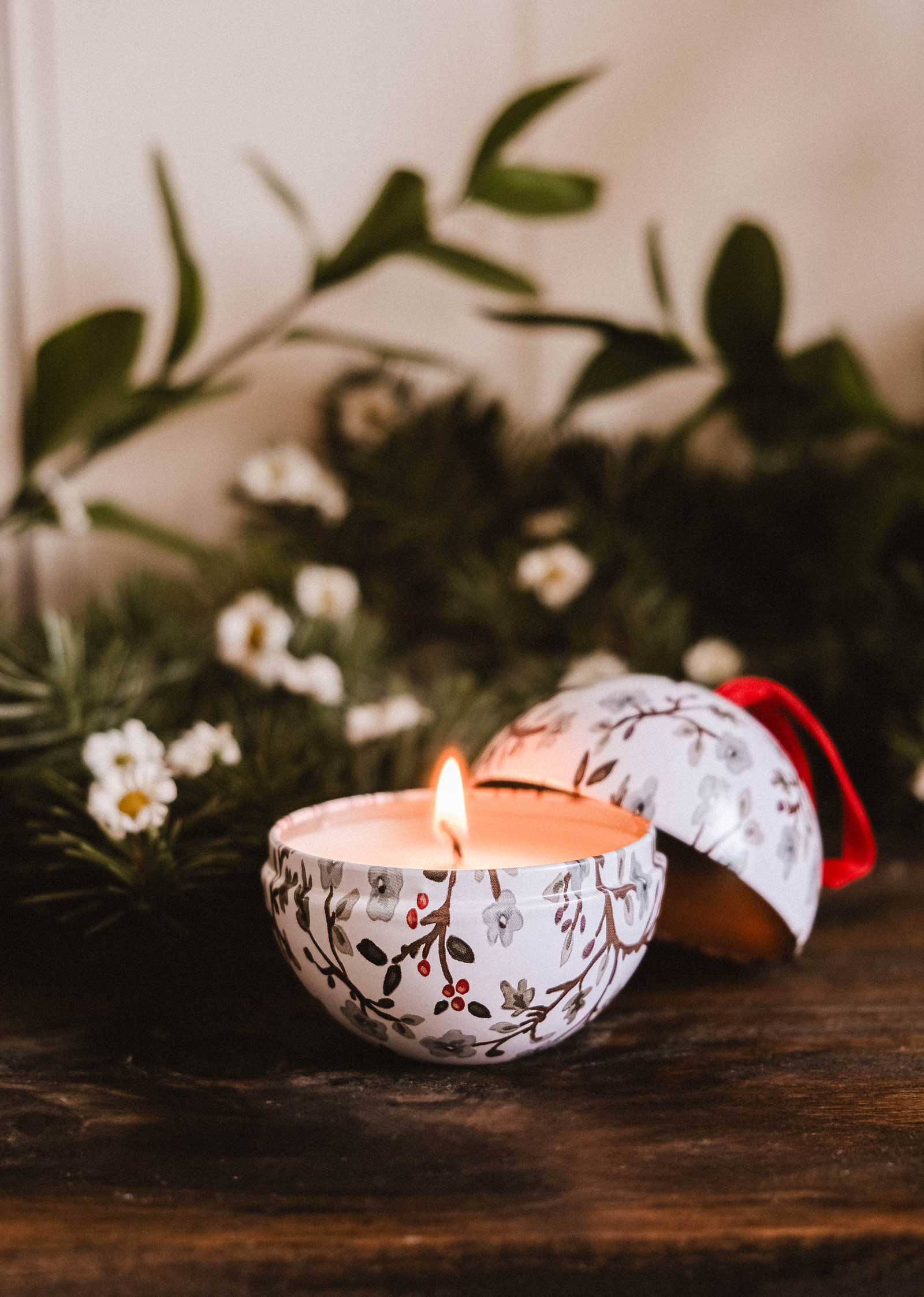 The Winter Flower - Christmas Ornament Candle by Mimi & August glows in a round, floral-patterned tin on a wooden surface with greenery and white flowers. The reusable ornament lid rests nearby.