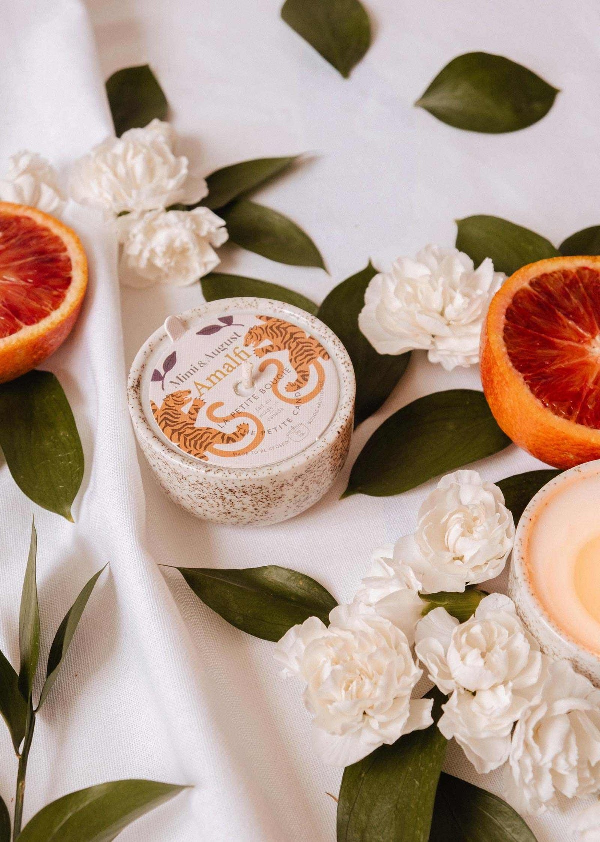 A round "Amalfi - Reusable Candle" by Mimi & August rests amid white flowers, tropical leaves, and blood orange slices on white fabric, capturing the essence of seaside holidays.