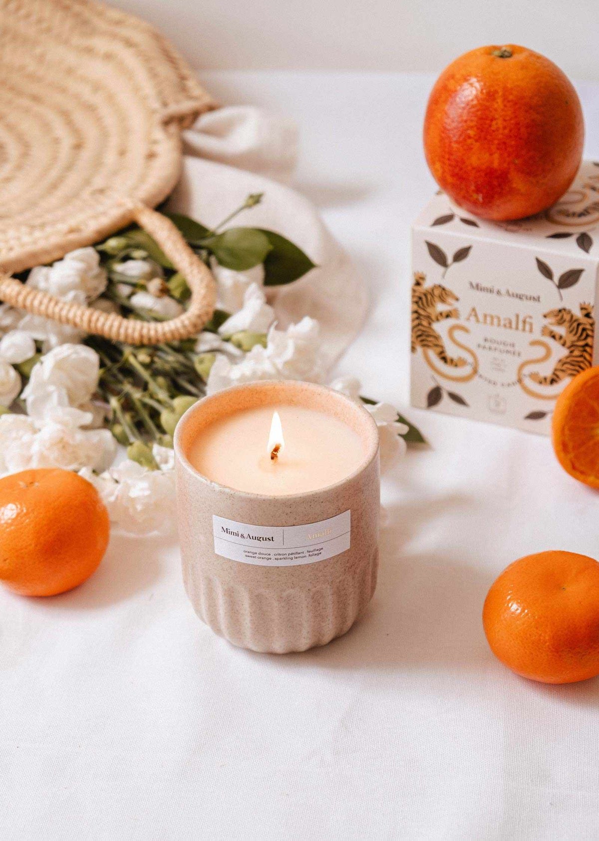 A lit Mimi & August Amalfi - Reusable Candle in a beige container rests on a white surface, surrounded by oranges, white flowers, tropical leaves, a woven bag, and an “Amalfi” box—evoking the spirit of seaside holidays.