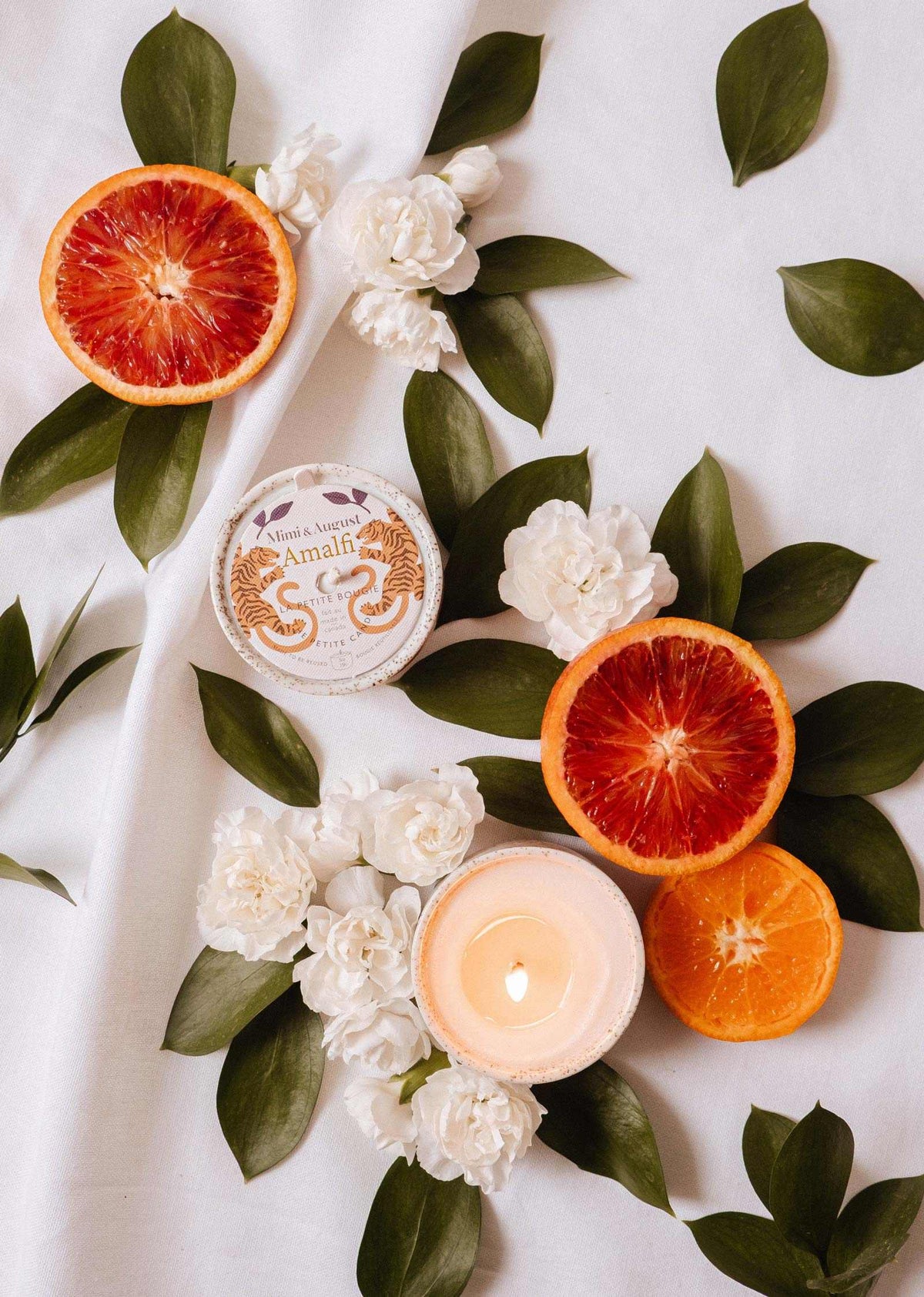 Sliced blood oranges, white flowers, tropical leaves, and the Mimi & August Amalfi - Reusable Candle are beautifully arranged on white fabric, evoking seaside holiday memories.