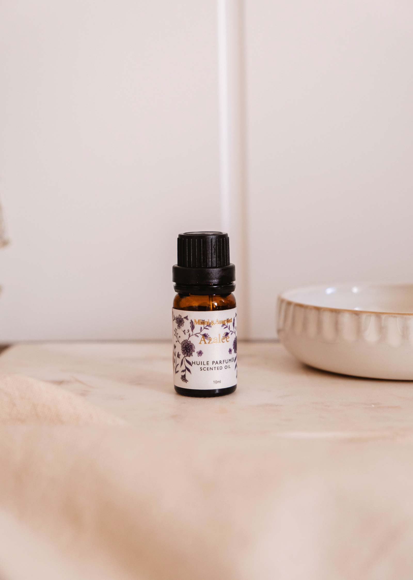A small amber glass bottle of Mimi & August Scented Oil - Azalée with a black cap and floral label rests on a light surface beside a ceramic dish.
