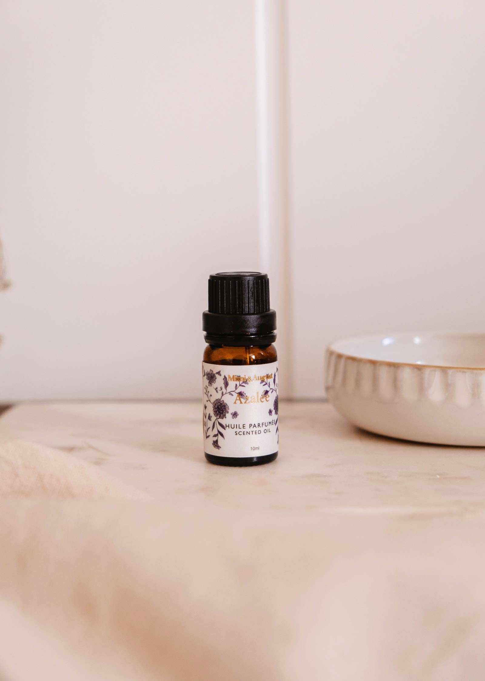 A small amber glass bottle of Mimi & August Scented Oil - Azalée with a black cap and floral label rests on a light surface beside a ceramic dish.