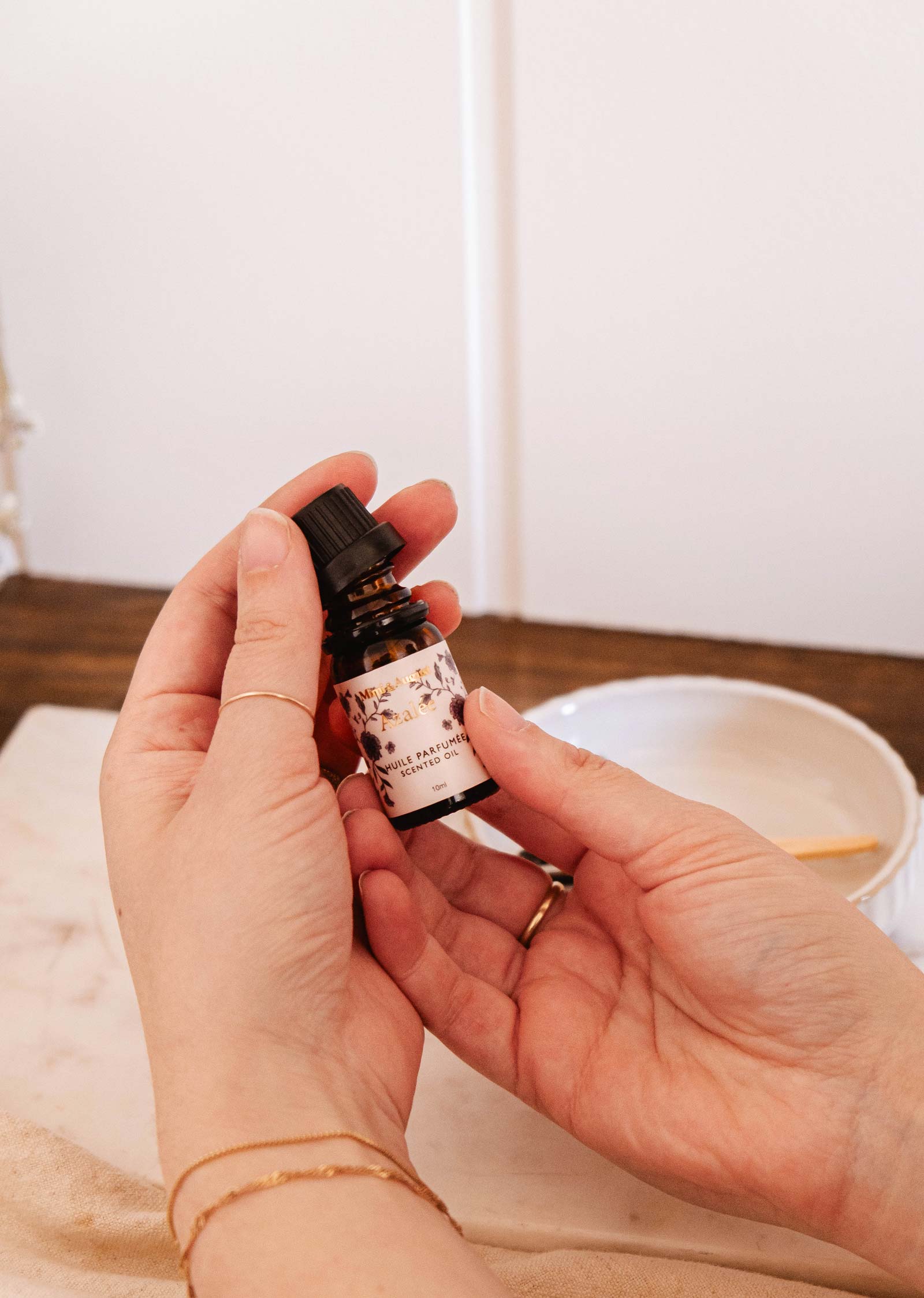 A person holds a small bottle of Mimi & August Scented Oil - Azalée with a floral label above a white ceramic dish on a wooden surface.
