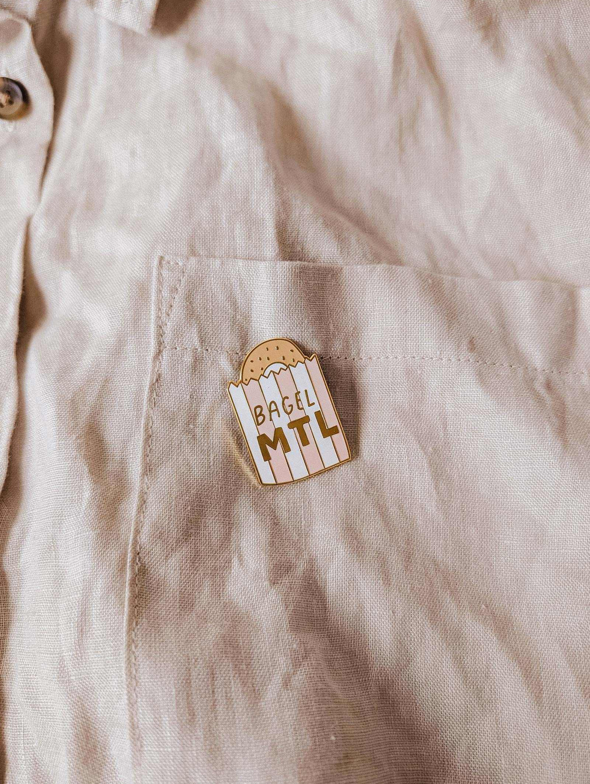 La poche d’une chemise beige met en valeur l’épinglette Bagel MTL de Mimi & August, illustrée d’un bagel et du texte « BAGEL MTL » — l’accessoire parfait pour les fans de bagels montréalais et les amoureux de bonne bouffe.