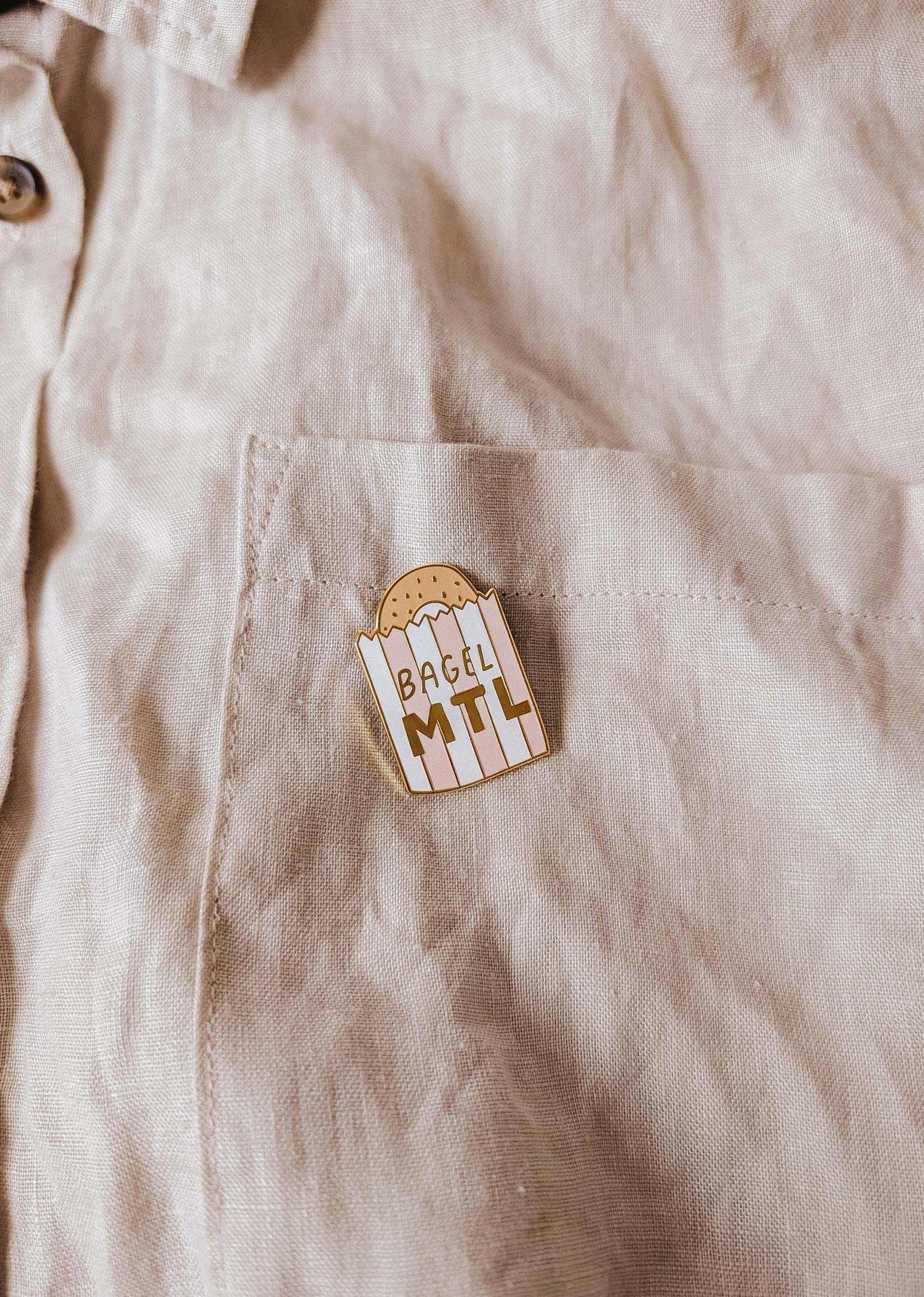 La poche d’une chemise beige met en valeur l’épinglette Bagel MTL de Mimi & August, illustrée d’un bagel et du texte « BAGEL MTL » — l’accessoire parfait pour les fans de bagels montréalais et les amoureux de bonne bouffe.