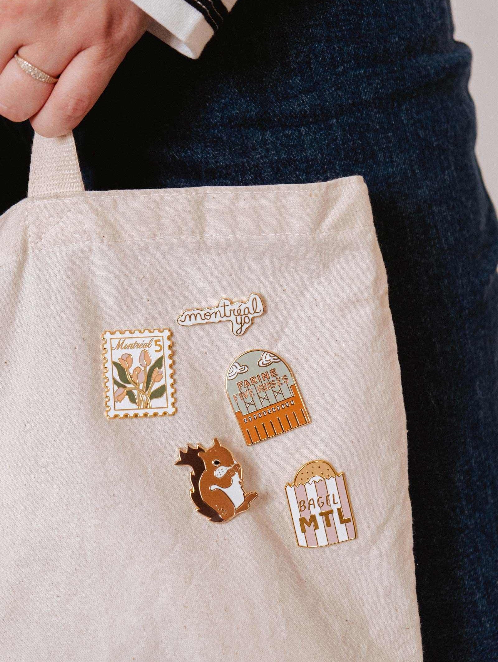 A close-up of a canvas tote bag featuring four Flour & Roses Factory Enamel Pins by Mimi & August, held by a person in denim with a ring.