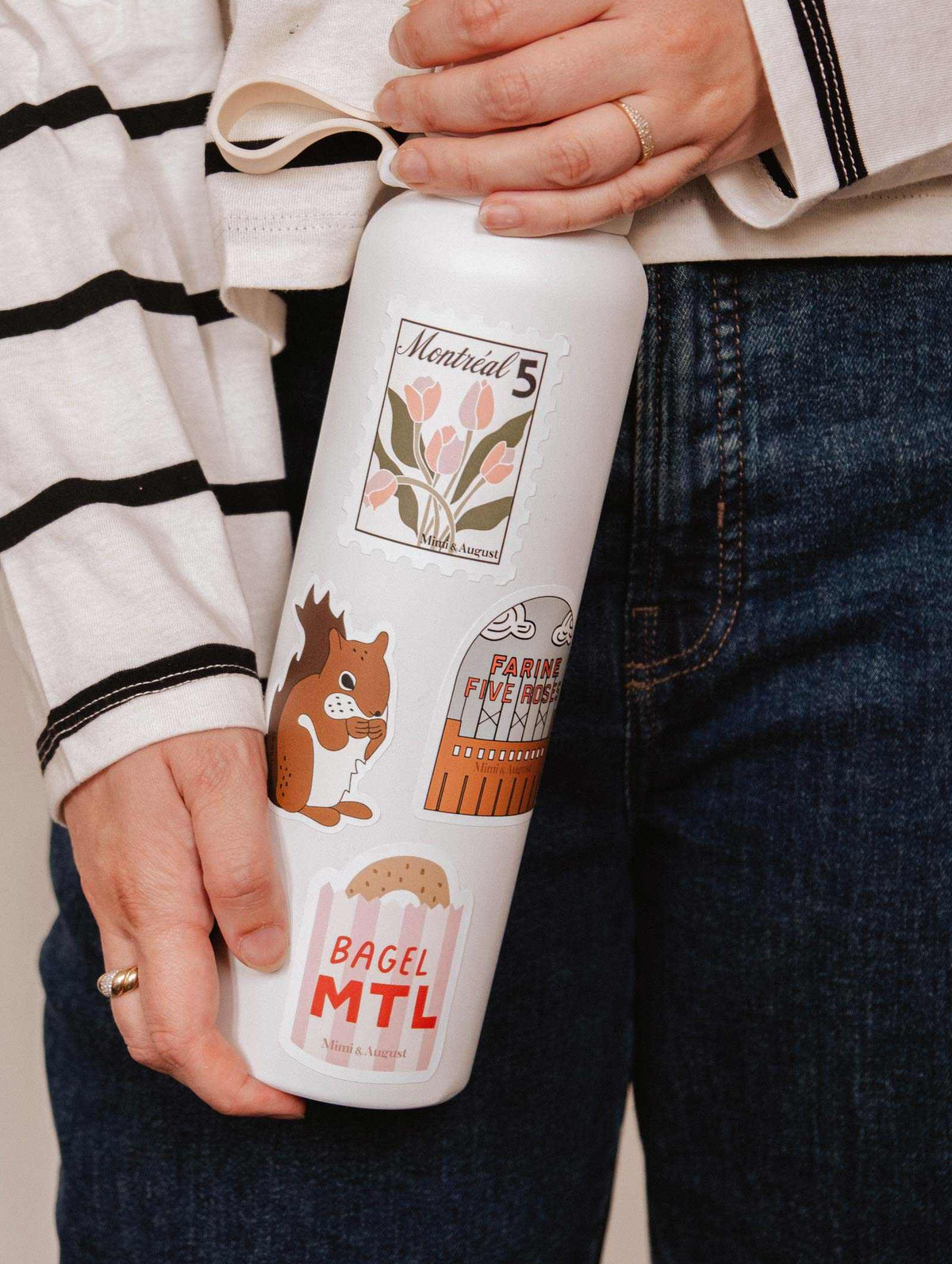 A person in jeans and a striped shirt holds a white water bottle decorated with Mimi & August’s Flour & Roses Factory vinyl sticker—an illustrated Montréal landmark, perfect for architecture lovers.