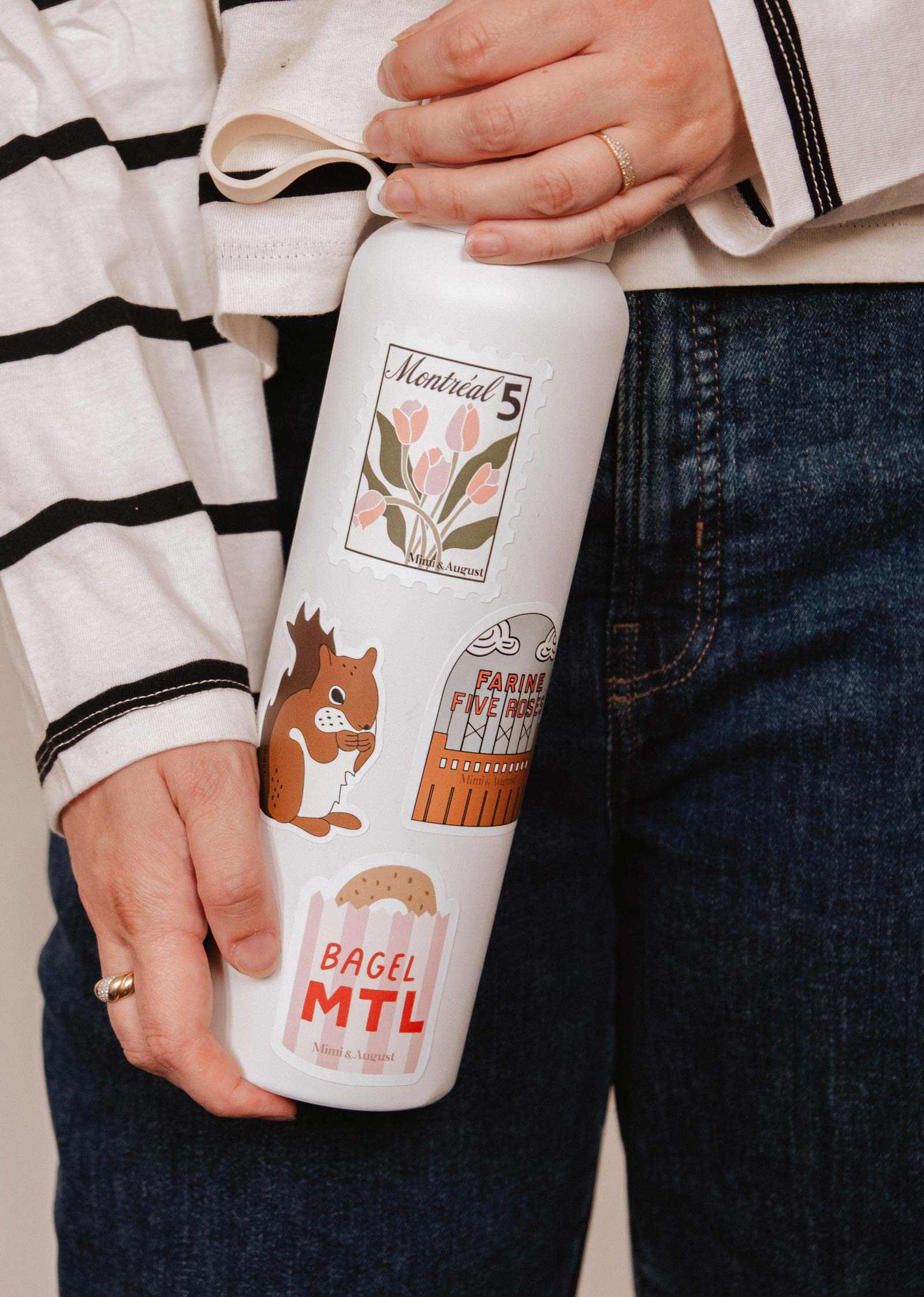 A person in a striped shirt and jeans holds a white water bottle adorned with Mimi & August vinyl stickers, including flowers, a squirrel, a sign, and the iconic Bagel MTL - Vinyl Sticker—a true Montréal emblem.