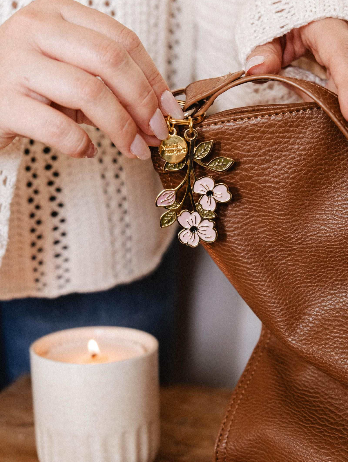 Une personne ajoute le Porte-clés Branche en Fleurs de Mimi & August à un sac en cuir brun, avec une bougie allumée à proximité sur une surface en bois.