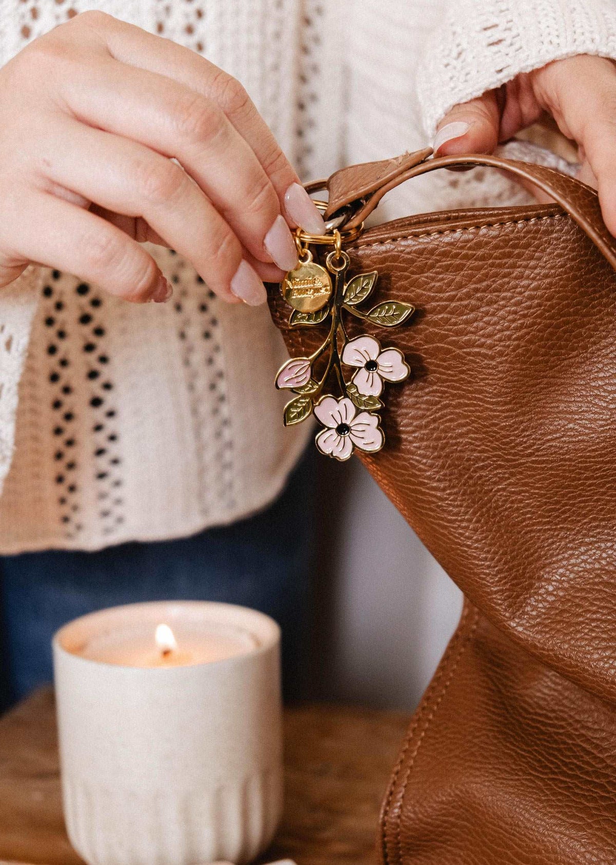 Someone adds the Mimi & August Blossom Branch Keychain to a brown leather bag, with a lit candle nearby on a wooden surface.