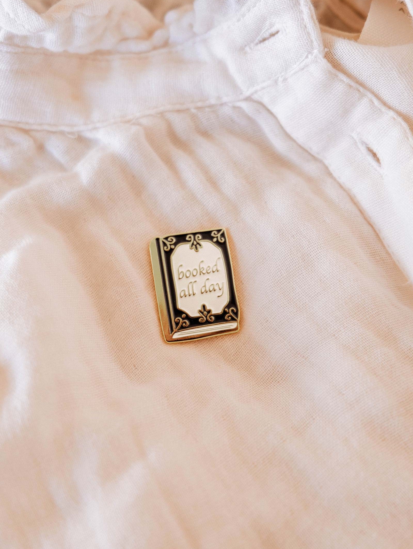 The Booked All Day Enamel Pin by Mimi & August features "booked all day" in cursive—an ideal bookworm gift and a stylish bookish accent for any white shirt or tote.