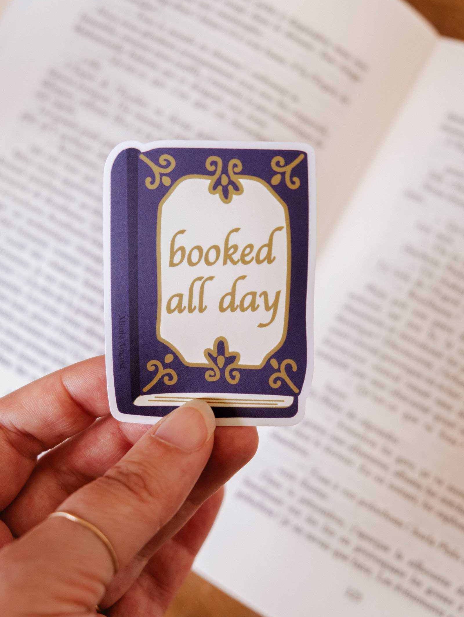 A hand holds the Mimi & August "Booked All Day" vinyl sticker, shaped like a book and perfect as a reading journal accessory.