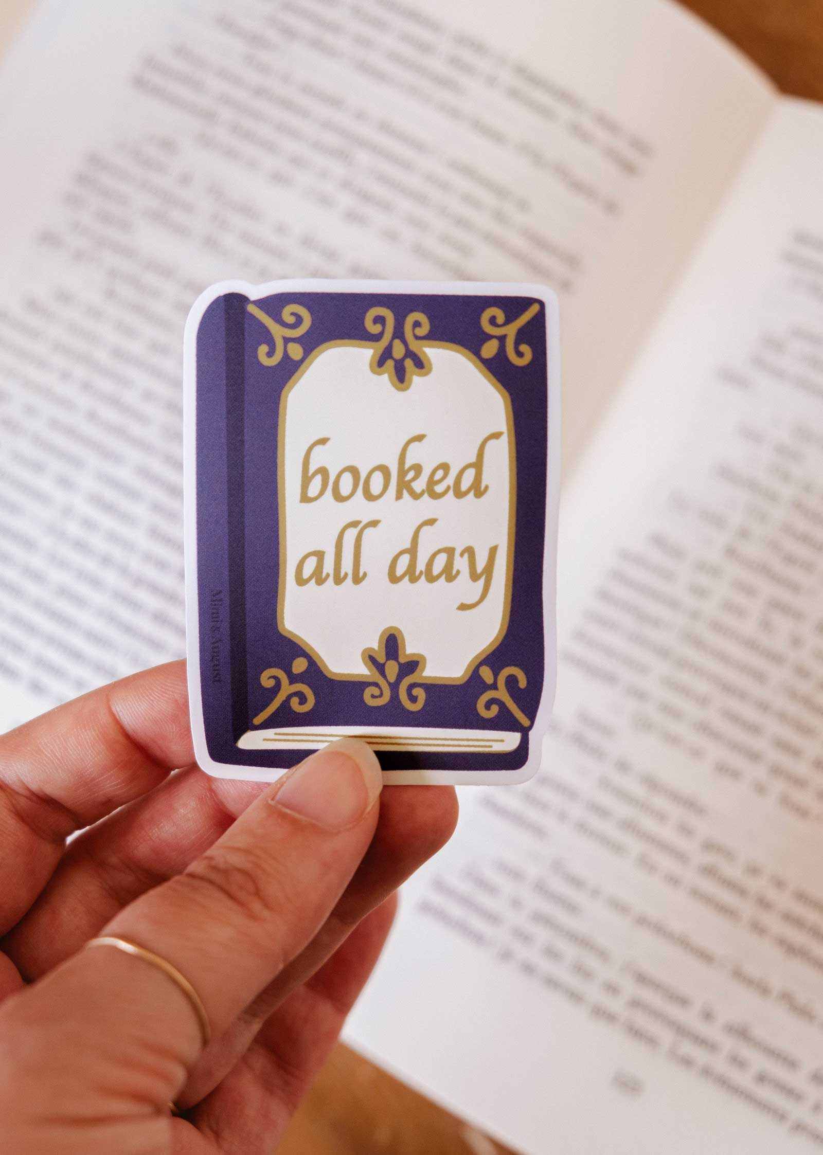 A hand holds the Mimi & August "Booked All Day" vinyl sticker, shaped like a book and perfect as a reading journal accessory.