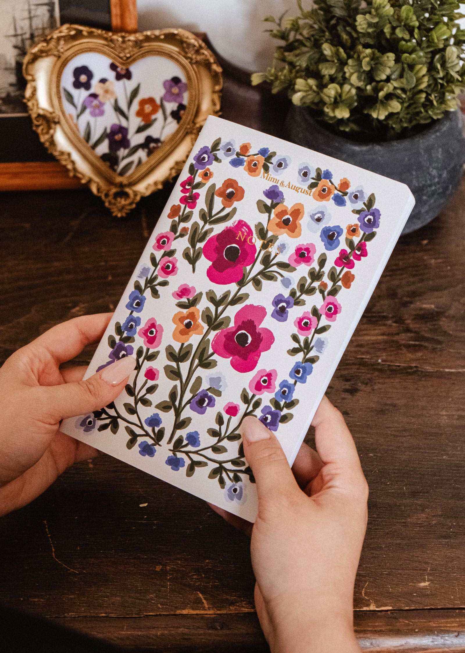 Two hands hold the Bouquet floral notebook by Mimi & August, with hand-painted florals, at a wooden desk with a plant and framed art.