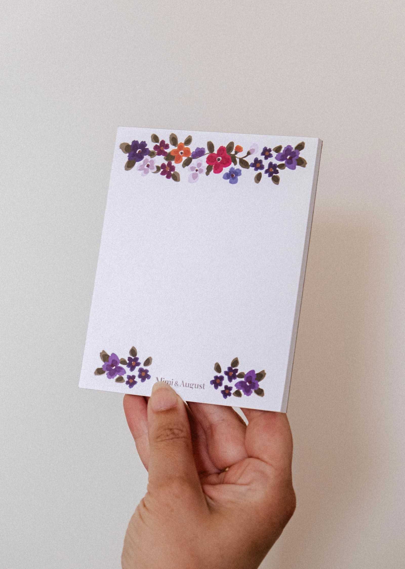 A hand holds the Bouquet floral notepad by Mimi & August, with purple and red blooms at the top and bottom, on a plain background.