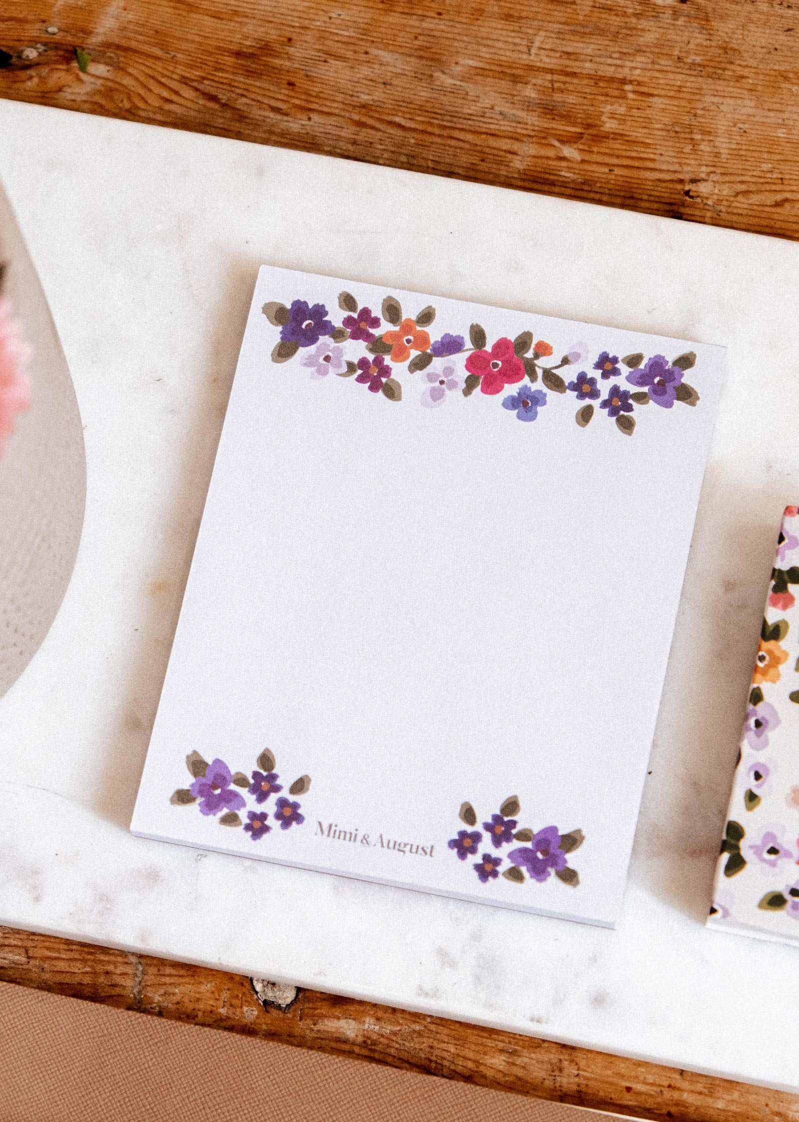 Bouquet floral notepad by Mimi & August, with hand-painted florals at the top and bottom, on a marble and wooden surface.