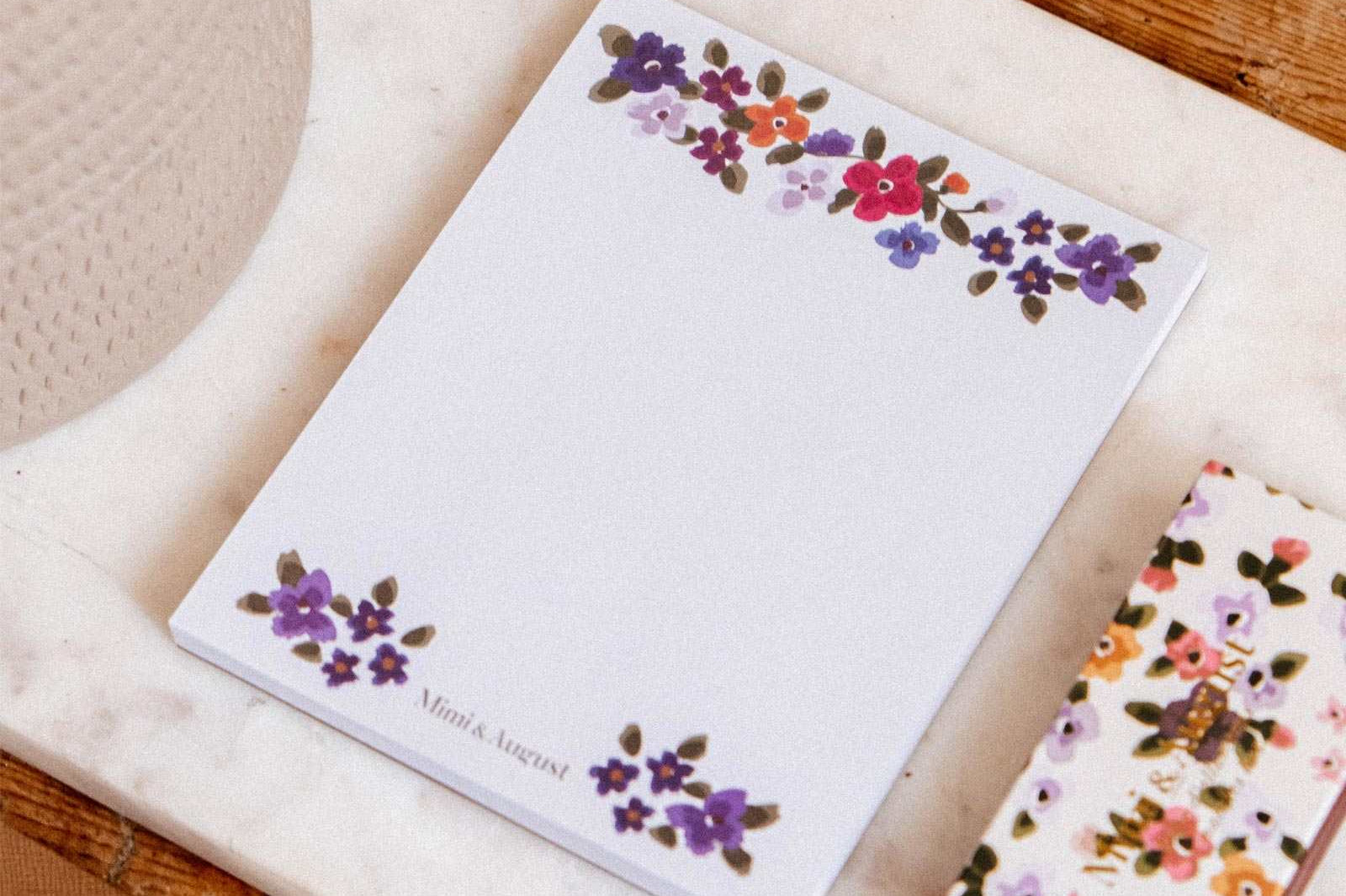 Bouquet floral notepad by Mimi & August with a matching pen on a marble surface beside a potted plant and wooden table.