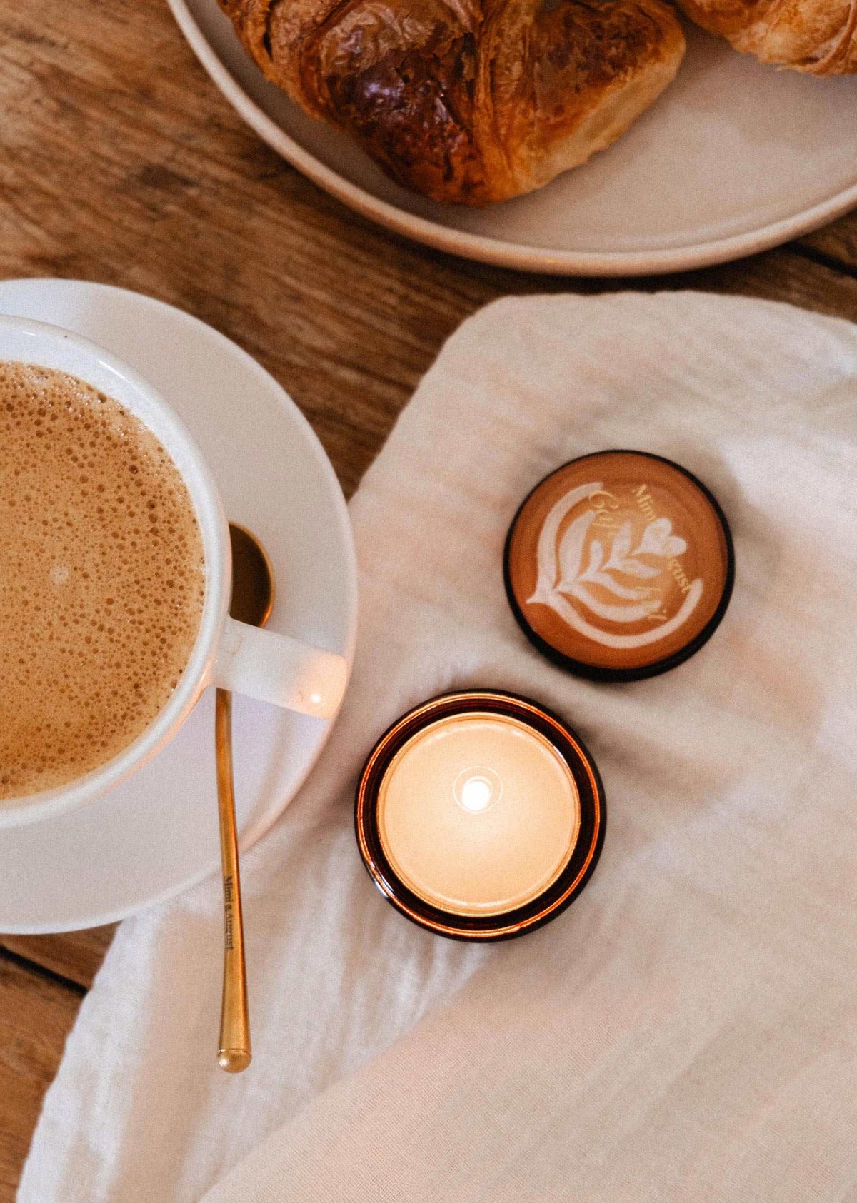 A cup of coffee, a Mimi & August Café au lait – 2 oz Soy Candle lit, and a croissant on a plate sit on a wooden table topped with a white cloth.