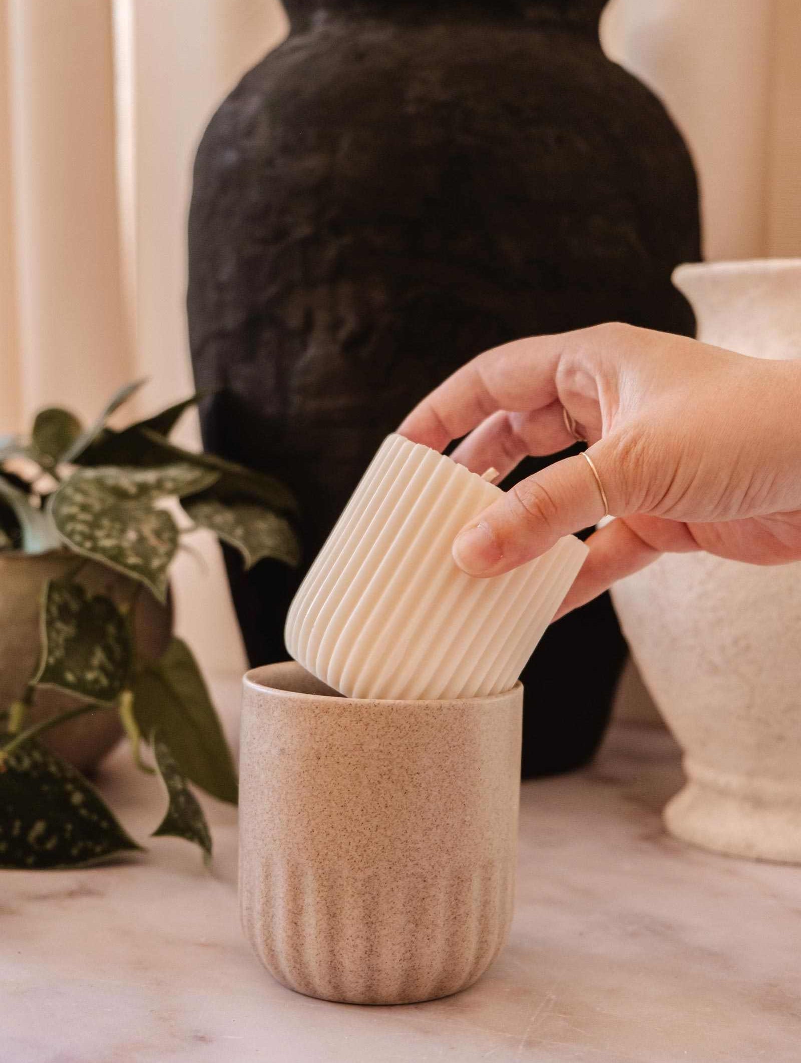 Une main dépose la Recharge de bougie Cielo de Mimi & August, un petit lampion en cire de soya blanc et cannelé, dans une grande tasse beige texturée sur une surface en marbre, avec des vases et une plante en arrière-plan.