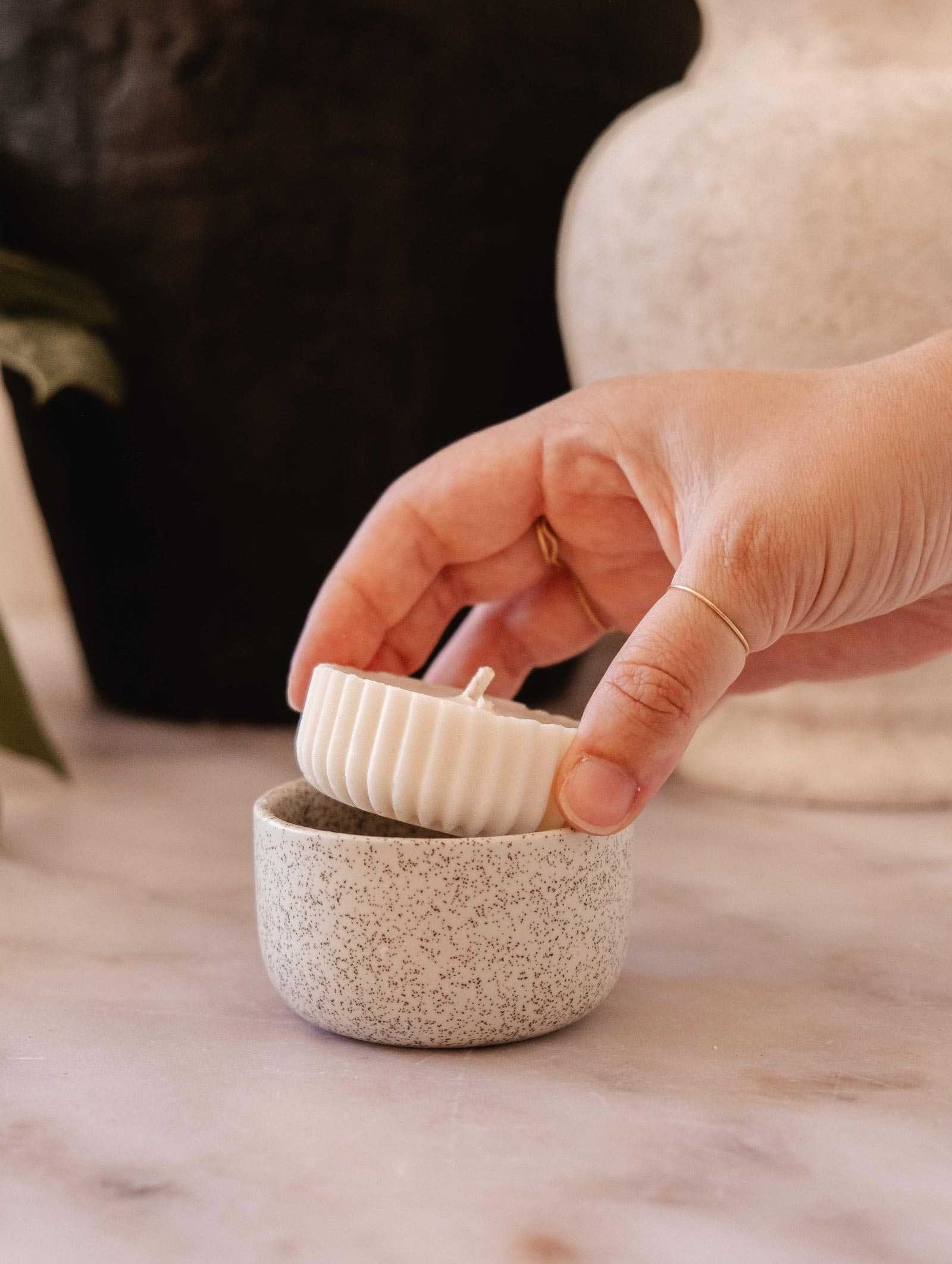 A hand lifts the lid of a small, round, speckled ceramic container on a marble surface to reveal the Mimi & August Candle Refill Alpes—an alpine-scented soy wax candle.