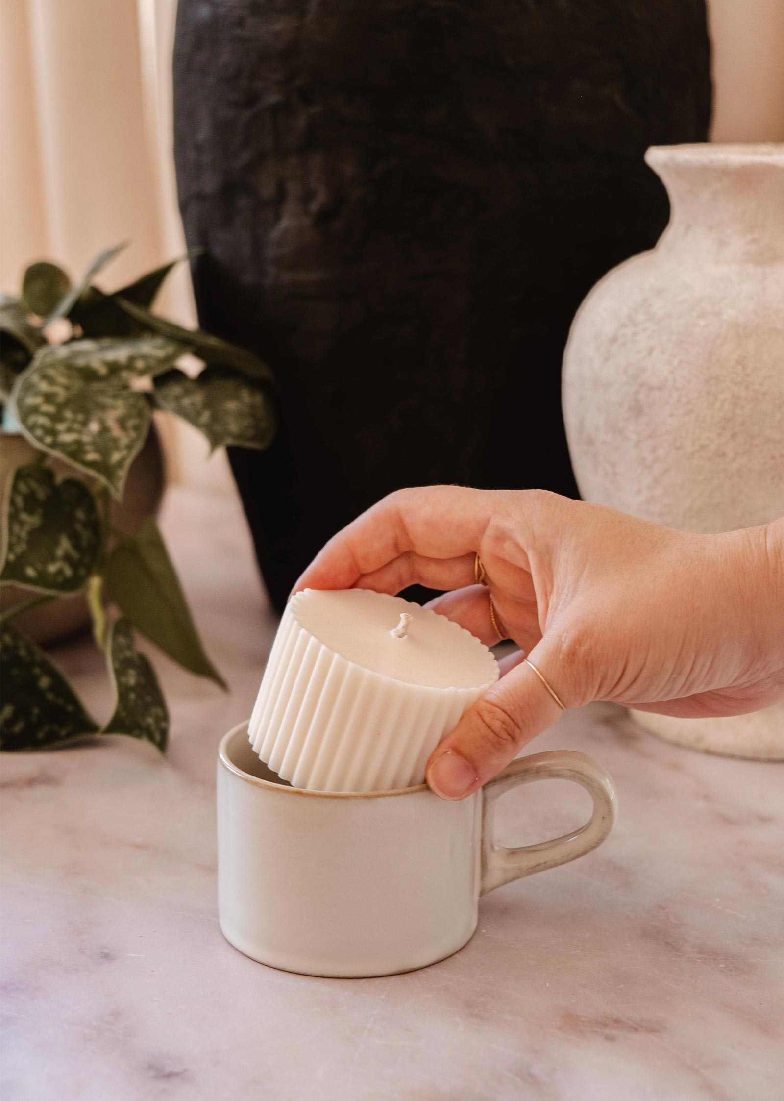 A hand places the Mimi & August Candle Refill Coco Mango, a white ribbed candle, into a white ceramic cup on a marble surface with plants and vases in the background.
