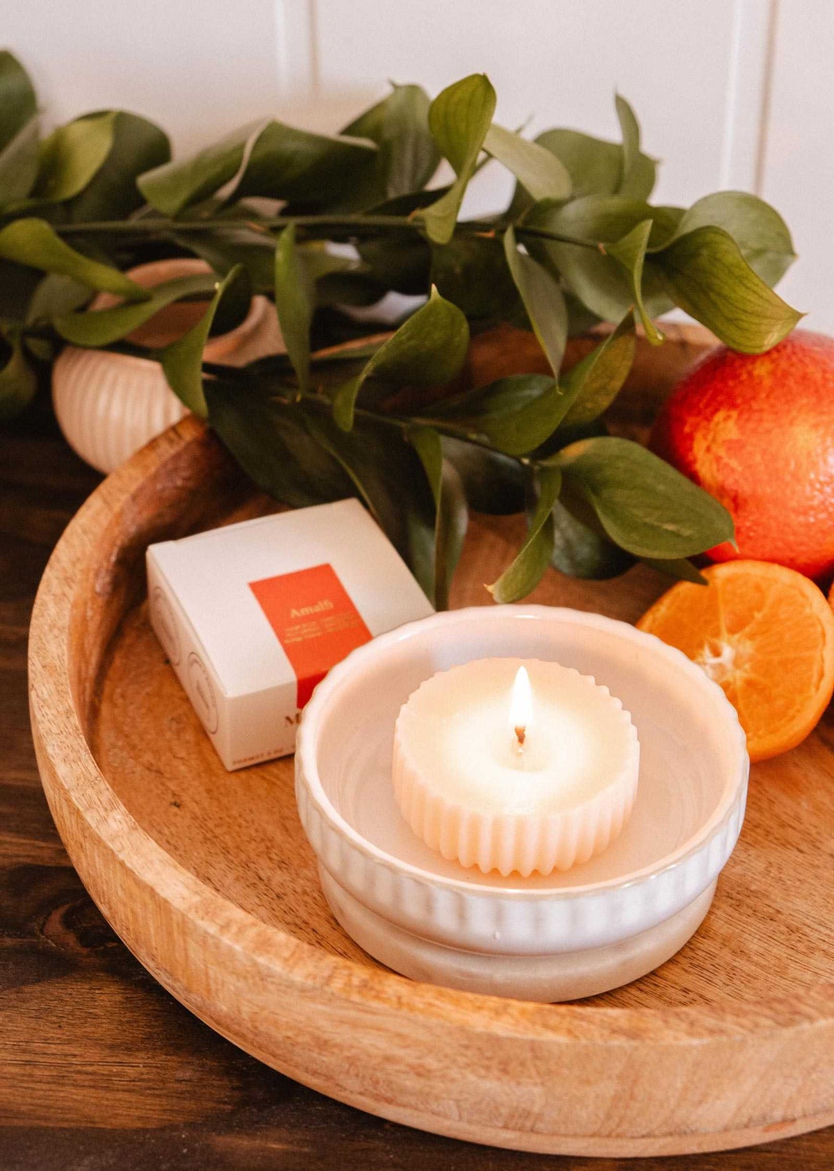La Recharge de bougie Amalfi de Mimi & August, présentée avec un plat blanc, sa boîte, de la verdure et des agrumes sur un plateau en bois, apporte une fragrance méditerranéenne fraîche à votre espace.