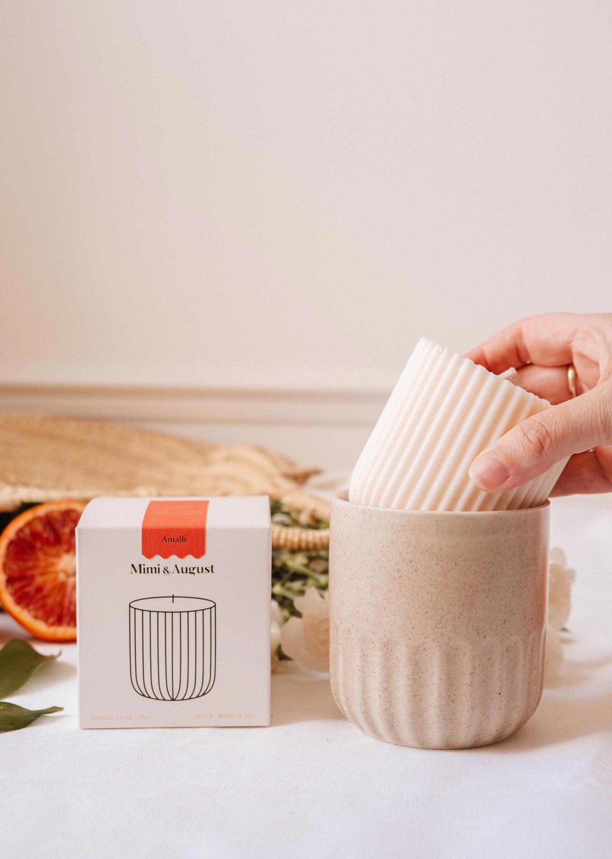 Une main dépose la Recharge de bougie Amalfi de Mimi & August, une bougie cannelée blanche en cire de soya naturelle, dans un bougeoir beige en céramique à côté de sa boîte, avec des tranches d'orange sanguine et de la verdure en arrière-plan.