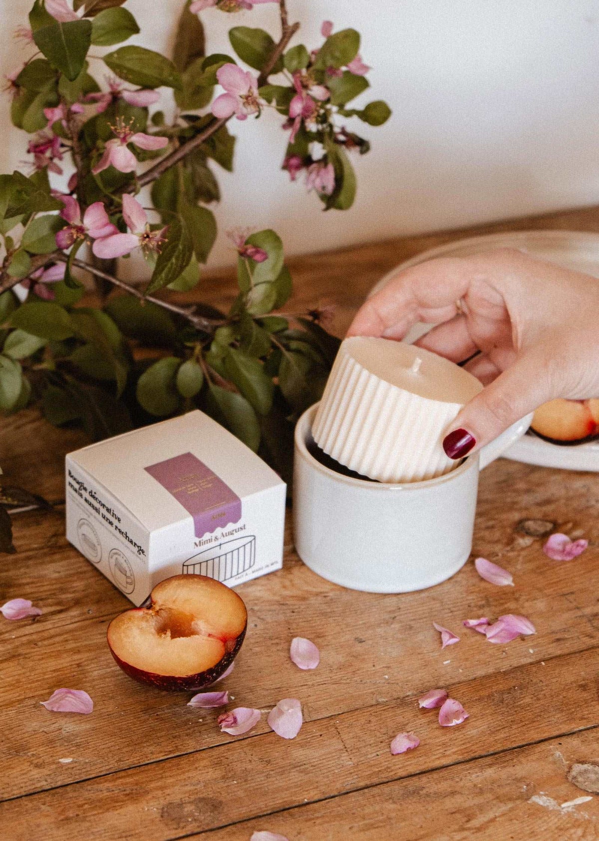 A hand places a white ribbed Candle Refill - Artea by Mimi & August into a white mug on a wooden table, surrounded by a halved plum, scattered flower petals, the Mimi & August product box, and flowering branches.