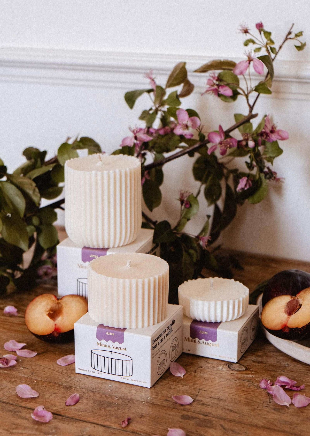 The Mimi & August Candle Refill - Artea, a ribbed white soy wax candle, is displayed on boxes with sliced plums and pink flowers atop a wooden surface, greenery in the background.